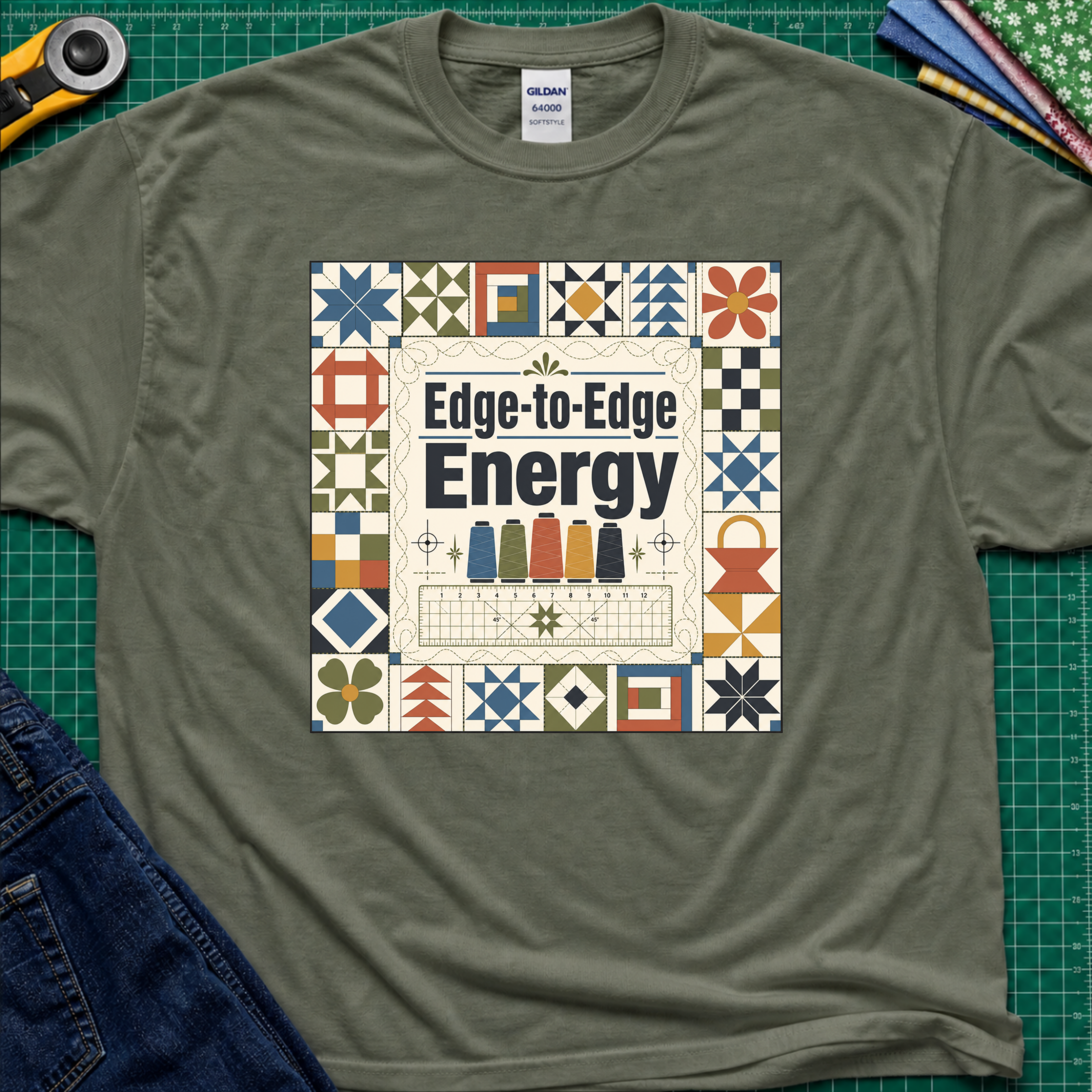 A military green Gildan t-shirt featuring the text 'Edge-to-Edge Energy' in the center, surrounded by quilt blocks. The t-shirt lays flat on a green cutting mat with a rotary cutter, folded fabric stack and blue jeans partially visible in the corners.