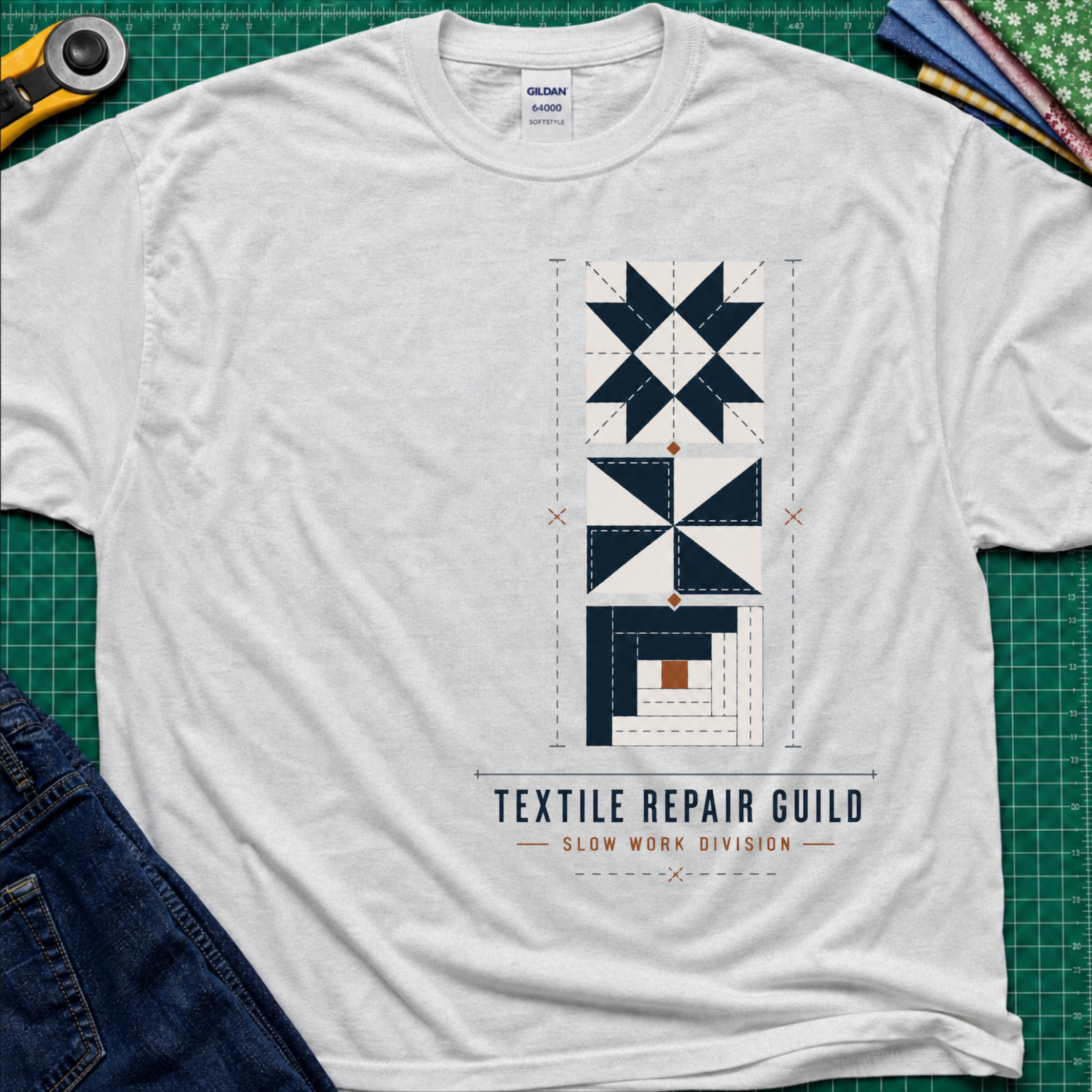 A light grey t-shirt featuring stacked quilt blocks in navy blue, white and tan with the text 'TEXTILE REPAIR GUILD' and 'SLOW WORK DIVISION', laying on a green cutting mat with a rotary cutter, fabric stack and jeans partially visible in the corners.