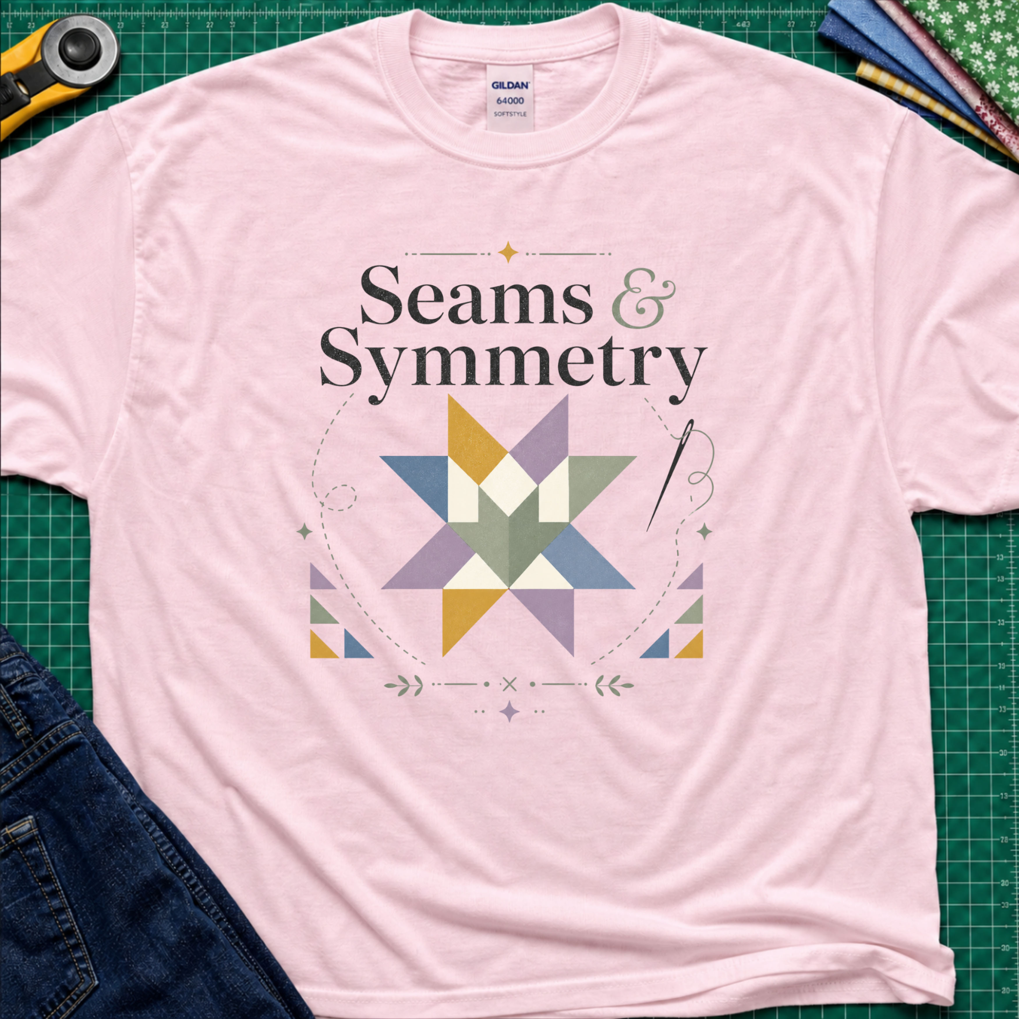 A light pink Gildan t-shirt with a 'Seams & Symmetry' graphic print with quilt block, needle and thread, laid flat on a green cutting mat with sewing props like a rotary cutter and denim jeans.