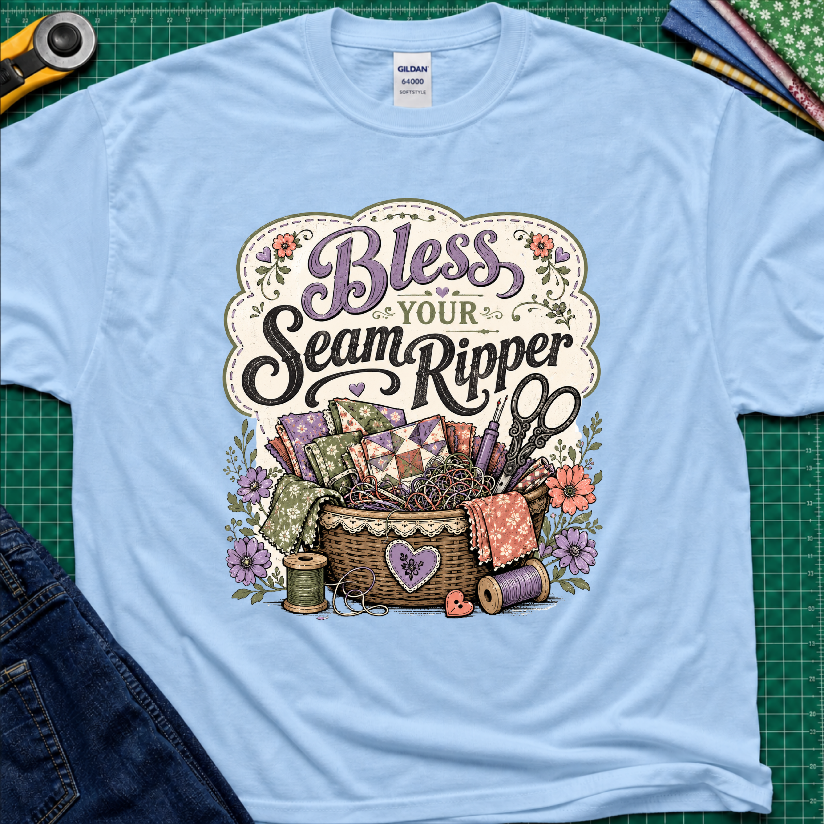A light blue t-shirt reading 'Bless Your Seam Ripper' above a basket of sewing supplies including thread, scissors, and fabric. The shirt lays on a green cutting mat with rotary cutter, fabric stack and jeans partially visible in the corners.