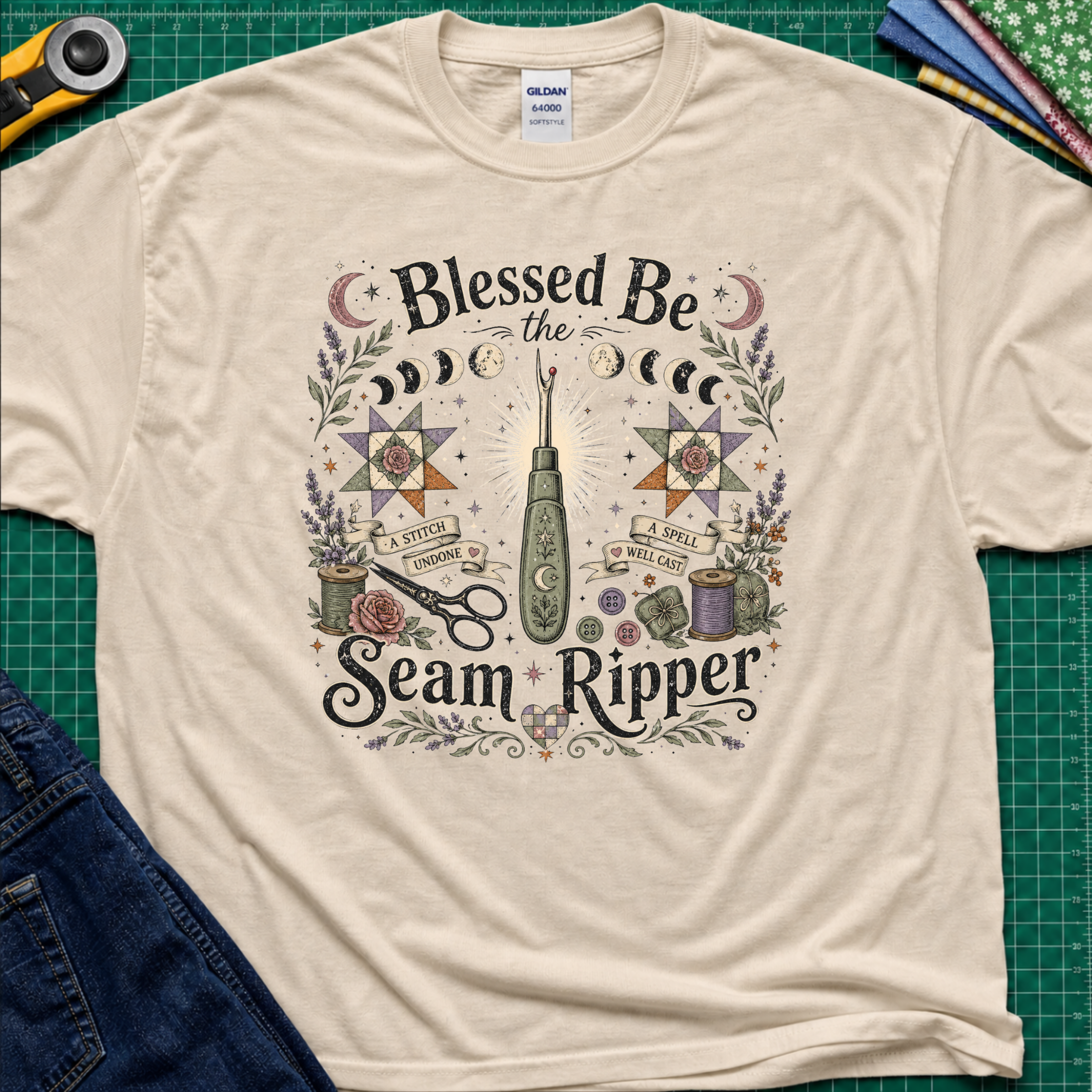 A dark cream t-shirt reading 'Blessed Be the Seam Ripper' surrounded by moon phases, quilt stars, thread spools with a back lit seam ripper in the middle, laying on a green cutting mat with rotary cutter, fabric stack and jeans partially.