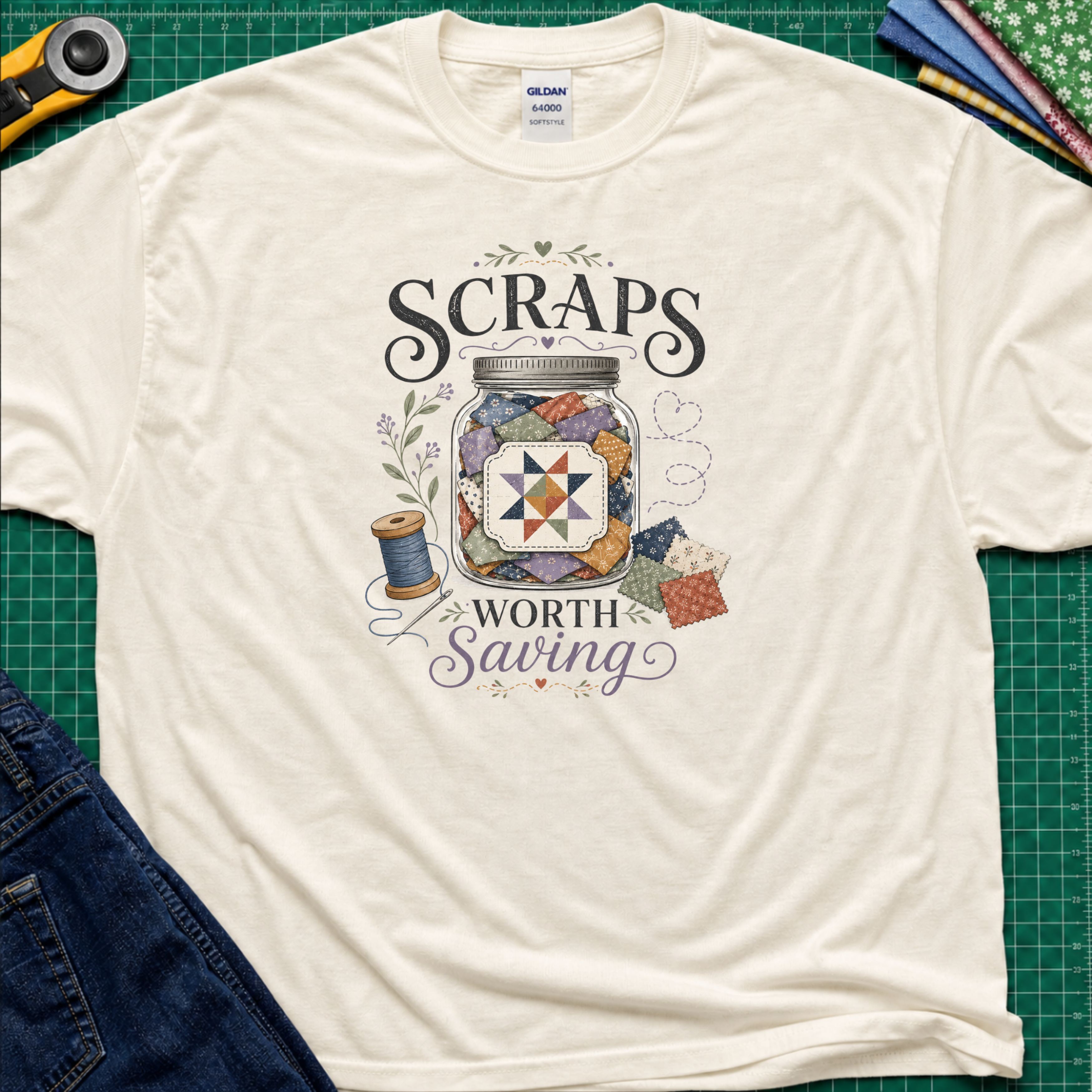 A cream t-shirt laying on a green cutting mat, reaing 'Scraps Worth Saving' around a jar filled with scraps, thread spool, and fabric pieces. A rotary cutter, scissors, and folded fabrics are visible around the shirt as styling props.