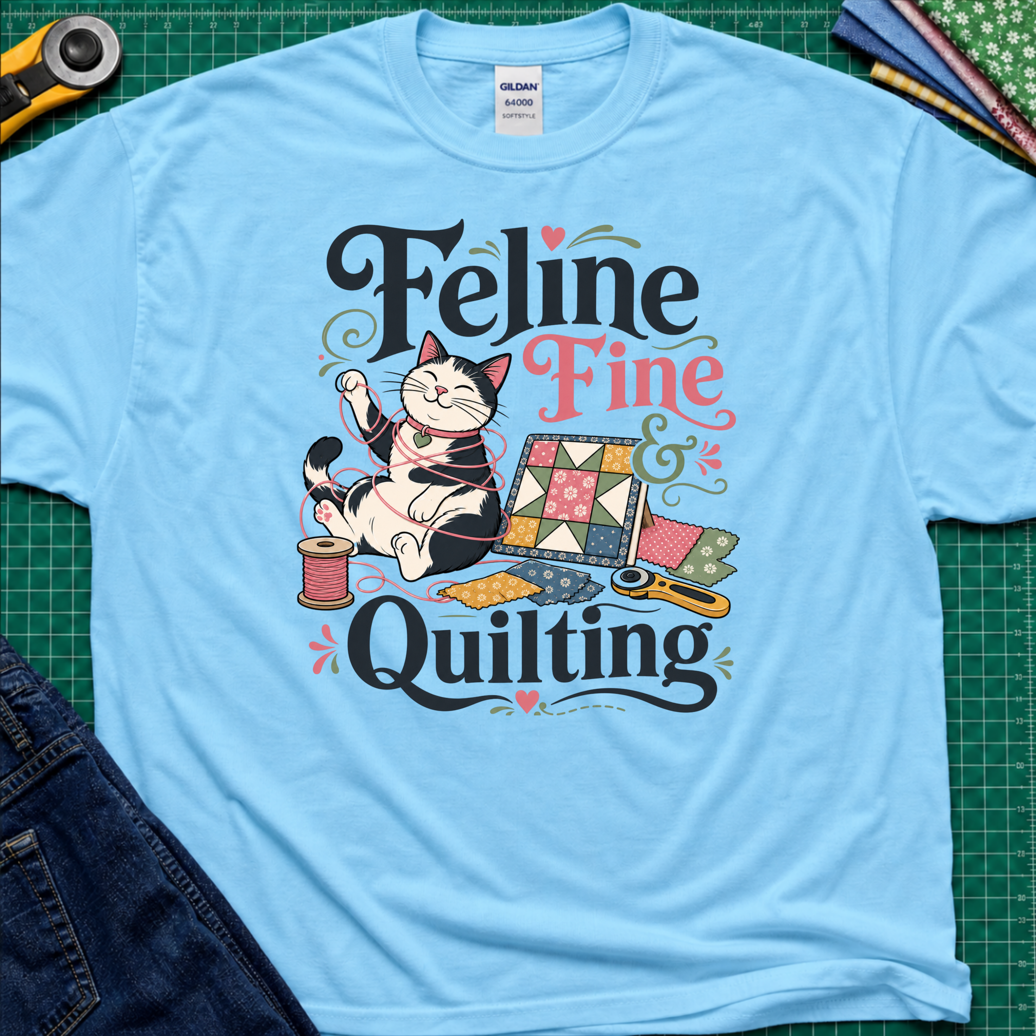 A sky blue Gildan t-shirt with a colorful graphic that reads 'Feline Fine Quilting' featuring a cartoon cat and quilting supplies, laid flat on a green cutting mat with crafting props and blue jeans partially visible.