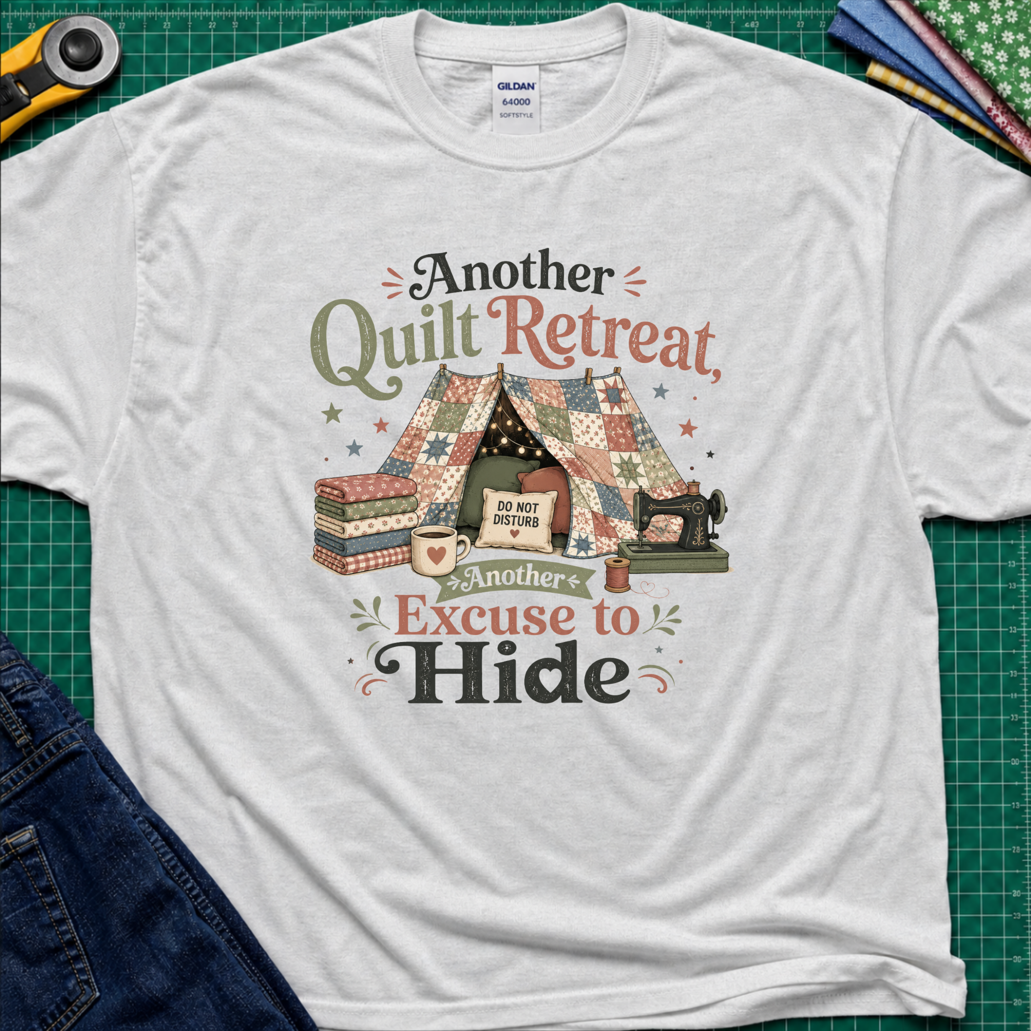 An ice grey Gildan t-shirt with a vintage-style quilted tent graphic and the text 'Another Quilt Retreat, Another Excuse to Hide' laid flat on a green cutting mat with sewing supplies.