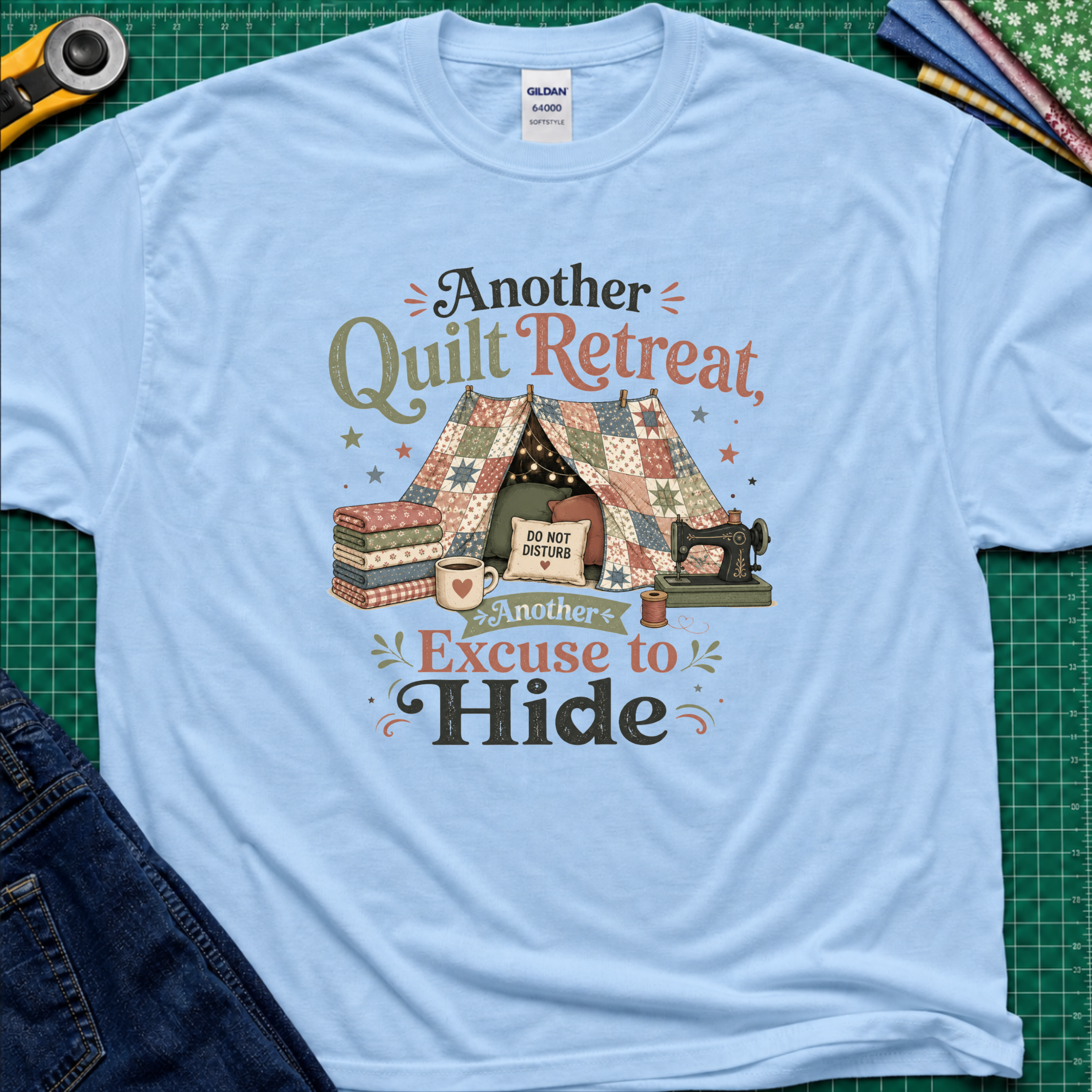 A light blue Gildan t-shirt with a vintage-style quilted tent graphic and the text 'Another Quilt Retreat, Another Excuse to Hide' laid flat on a green cutting mat with sewing supplies.