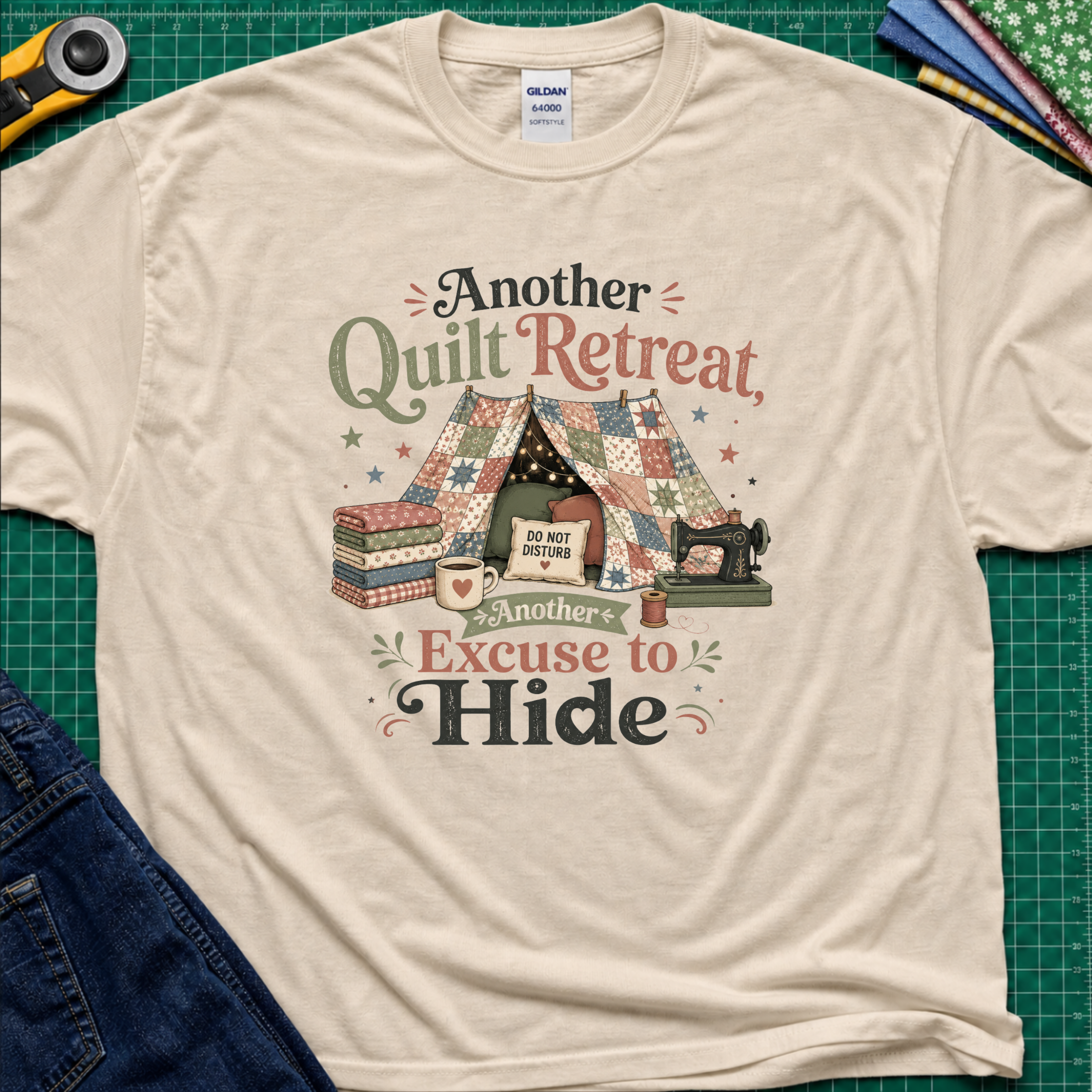 A dark cream Gildan t-shirt with a vintage-style quilted tent graphic and the text 'Another Quilt Retreat, Another Excuse to Hide' laid flat on a green cutting mat with sewing supplies.