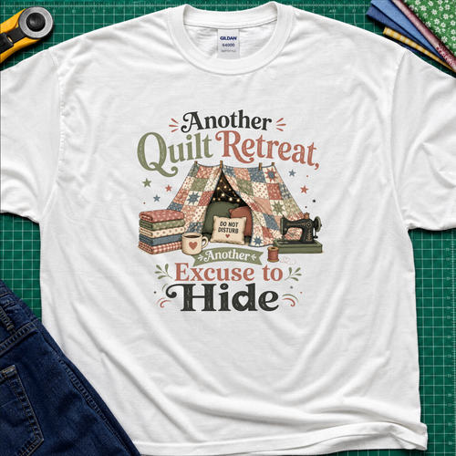 A white Gildan t-shirt with a vintage-style quilted tent graphic and the text 'Another Quilt Retreat, Another Excuse to Hide' laid flat on a green cutting mat with sewing supplies.