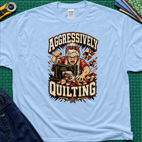 A light blue Gildan t-shirt laid flat on a green cutting mat, featuring a retro-style graphic of an elderly woman enthusiastically quilting with a sewing machine. The graphic includes the words 'AGGRESSIVELY QUILTING' in bold cream lettering.