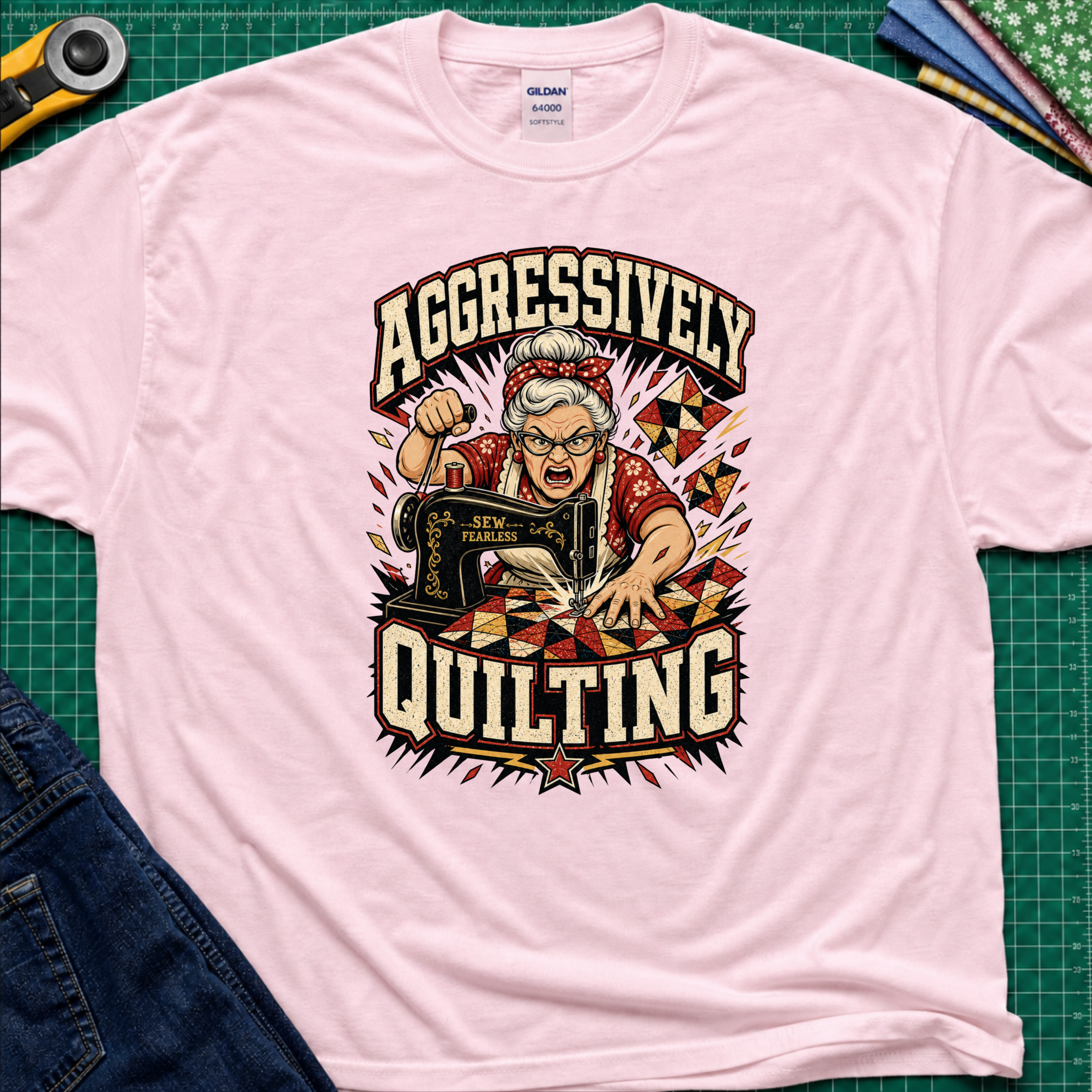 A light pink Gildan t-shirt laid flat on a green cutting mat, featuring a retro-style graphic of an elderly woman enthusiastically quilting with a sewing machine. The graphic includes the words 'AGGRESSIVELY QUILTING' in bold cream lettering.