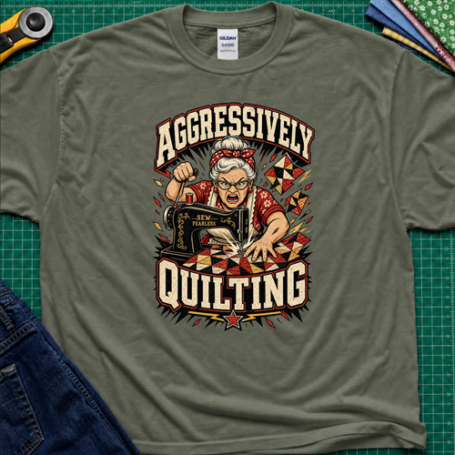 A military green Gildan t-shirt laid flat on a green cutting mat, featuring a retro-style graphic of an elderly woman enthusiastically quilting with a sewing machine. The graphic includes the words 'AGGRESSIVELY QUILTING' in bold cream lettering.