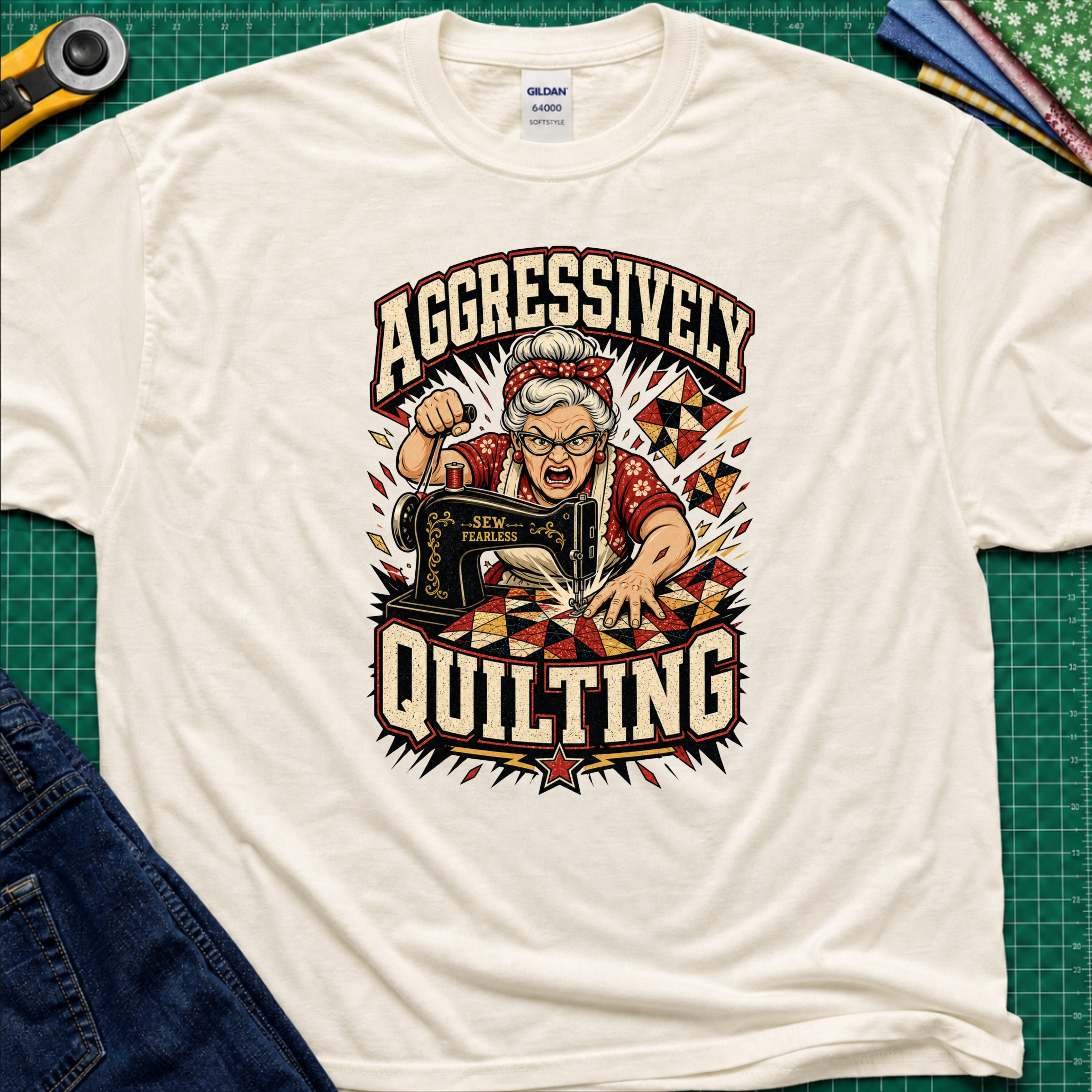 A light cream Gildan t-shirt laid flat on a green cutting mat, featuring a retro-style graphic of an elderly woman enthusiastically quilting with a sewing machine. The graphic includes the words 'AGGRESSIVELY QUILTING' in bold cream lettering.