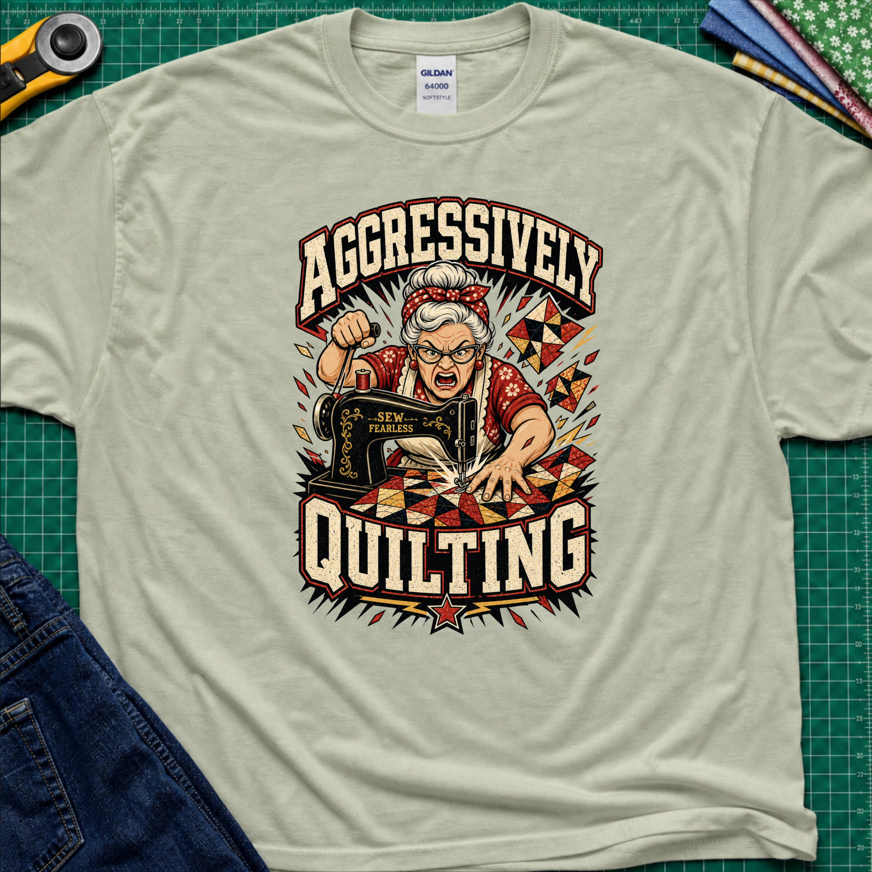 A light sage green Gildan t-shirt laid flat on a green cutting mat, featuring a retro-style graphic of an elderly woman enthusiastically quilting with a sewing machine. The graphic includes the words 'AGGRESSIVELY QUILTING' in bold cream lettering.