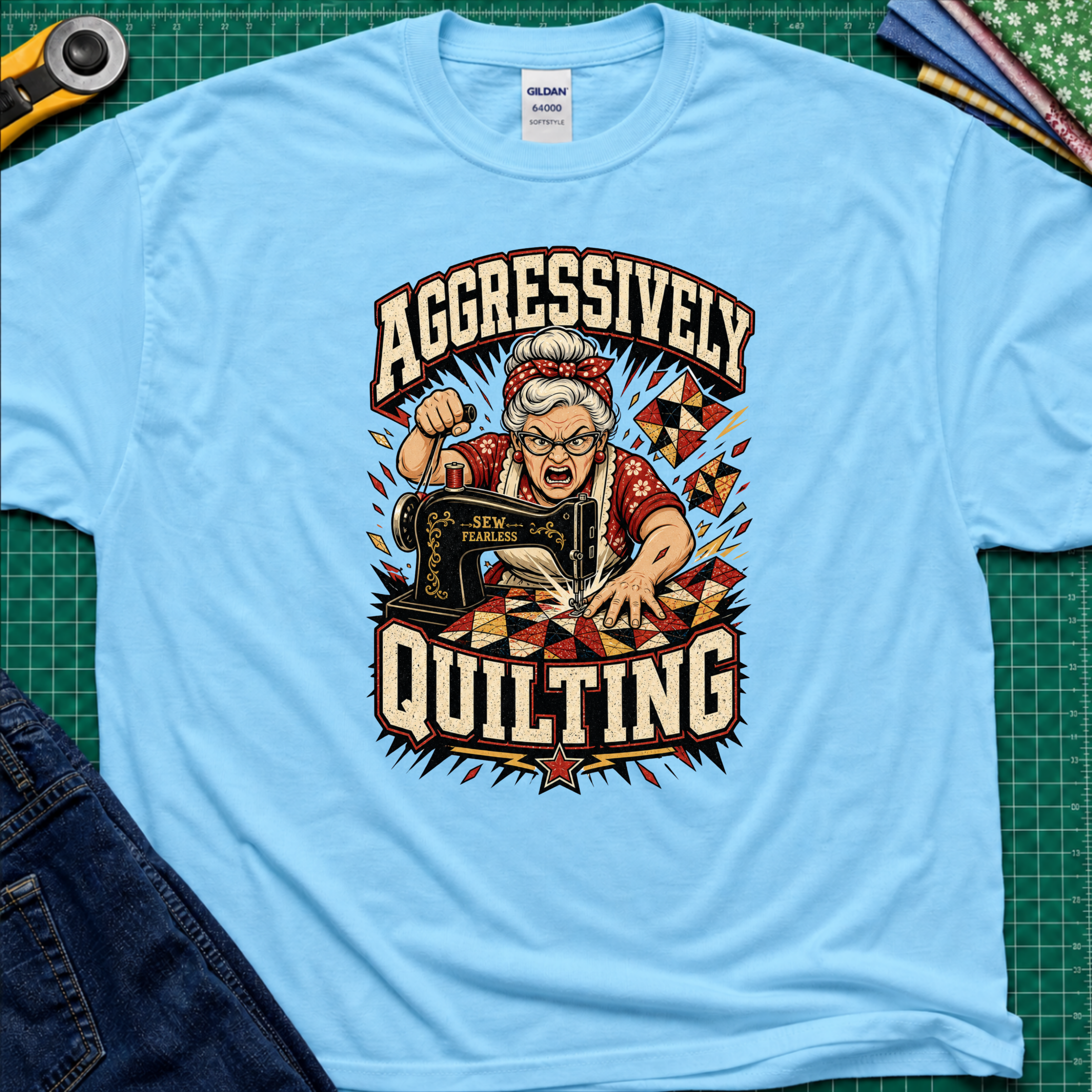 A sky blue Gildan t-shirt laid flat on a green cutting mat, featuring a retro-style graphic of an elderly woman enthusiastically quilting with a sewing machine. The graphic includes the words 'AGGRESSIVELY QUILTING' in bold cream lettering.