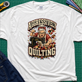 A white Gildan t-shirt laid flat on a green cutting mat, featuring a retro-style graphic of an elderly woman enthusiastically quilting with a sewing machine. The graphic includes the words 'AGGRESSIVELY QUILTING' in bold cream lettering.