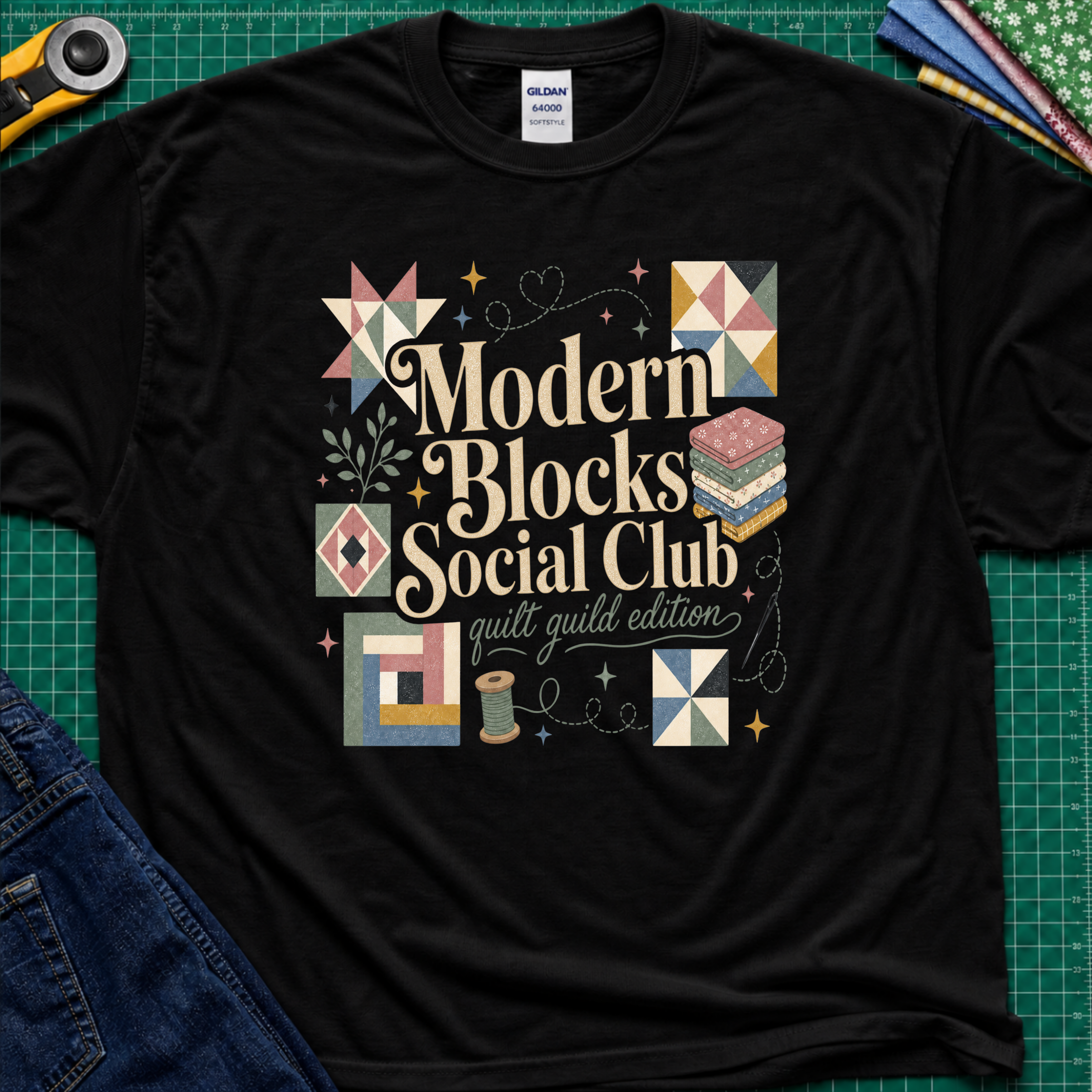 A black Gildan t-shirt with a colorful retro-style graphic reading 'Modern Blocks Social Club quilt guild edition' surrounded by quilt block illustrations. The shirt lays flat on a green cutting mat with a rotary cutter, fabric stack and blue jeans partially visible in the corners.
