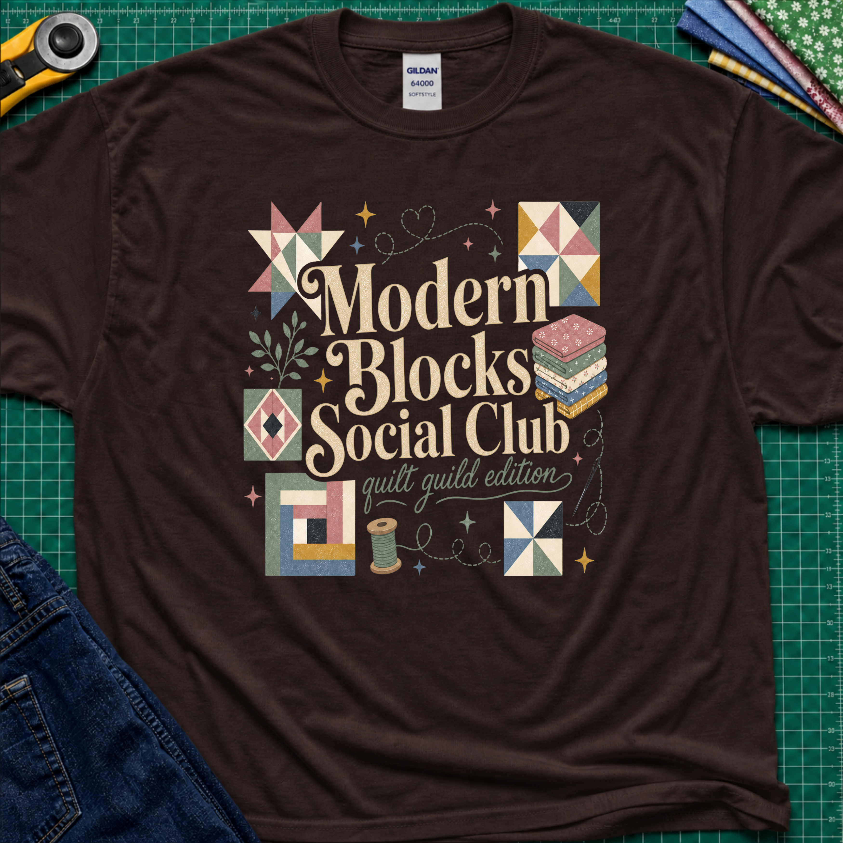 A dark chocolate Gildan t-shirt with a colorful retro-style graphic reading 'Modern Blocks Social Club quilt guild edition' surrounded by quilt block illustrations. The shirt lays flat on a green cutting mat with a rotary cutter, fabric stack and blue jeans partially visible in the corners.