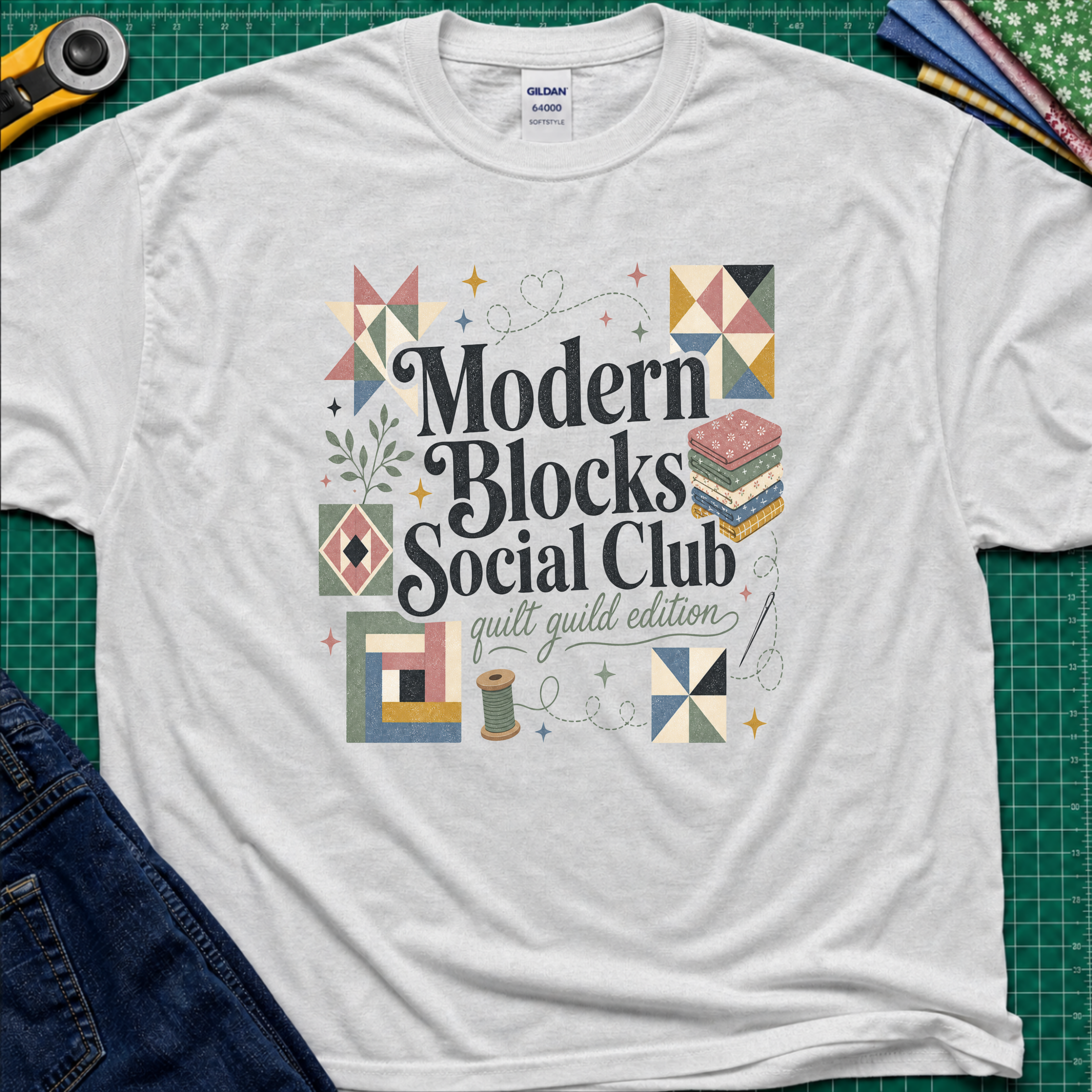 A light grey Gildan t-shirt with a colorful retro-style graphic reading 'Modern Blocks Social Club quilt guild edition' surrounded by quilt block illustrations. The shirt lays flat on a green cutting mat with a rotary cutter, fabric stack and blue jeans partially visible in the corners.