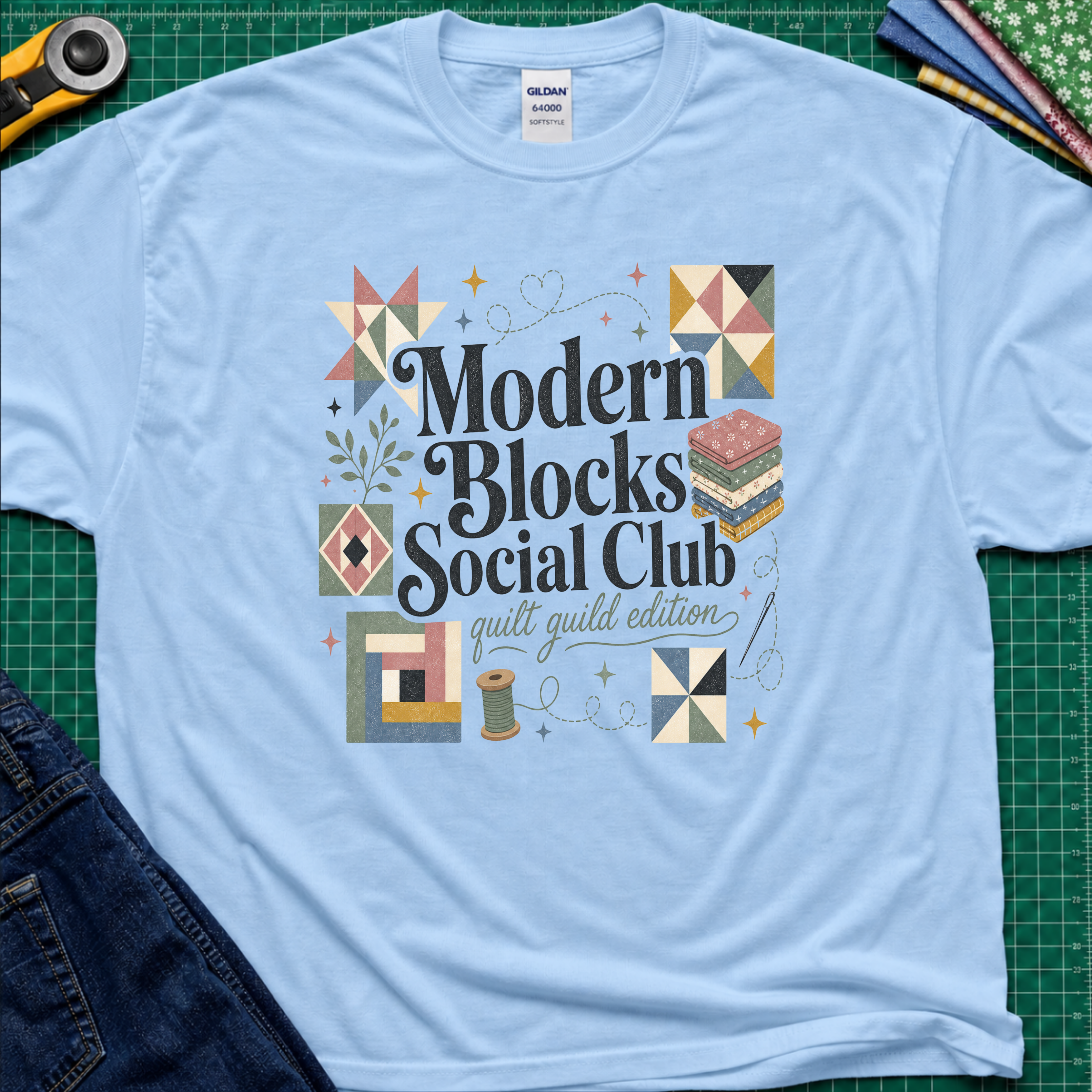 A light blue Gildan t-shirt with a colorful retro-style graphic reading 'Modern Blocks Social Club quilt guild edition' surrounded by quilt block illustrations. The shirt lays flat on a green cutting mat with a rotary cutter, fabric stack and blue jeans partially visible in the corners.