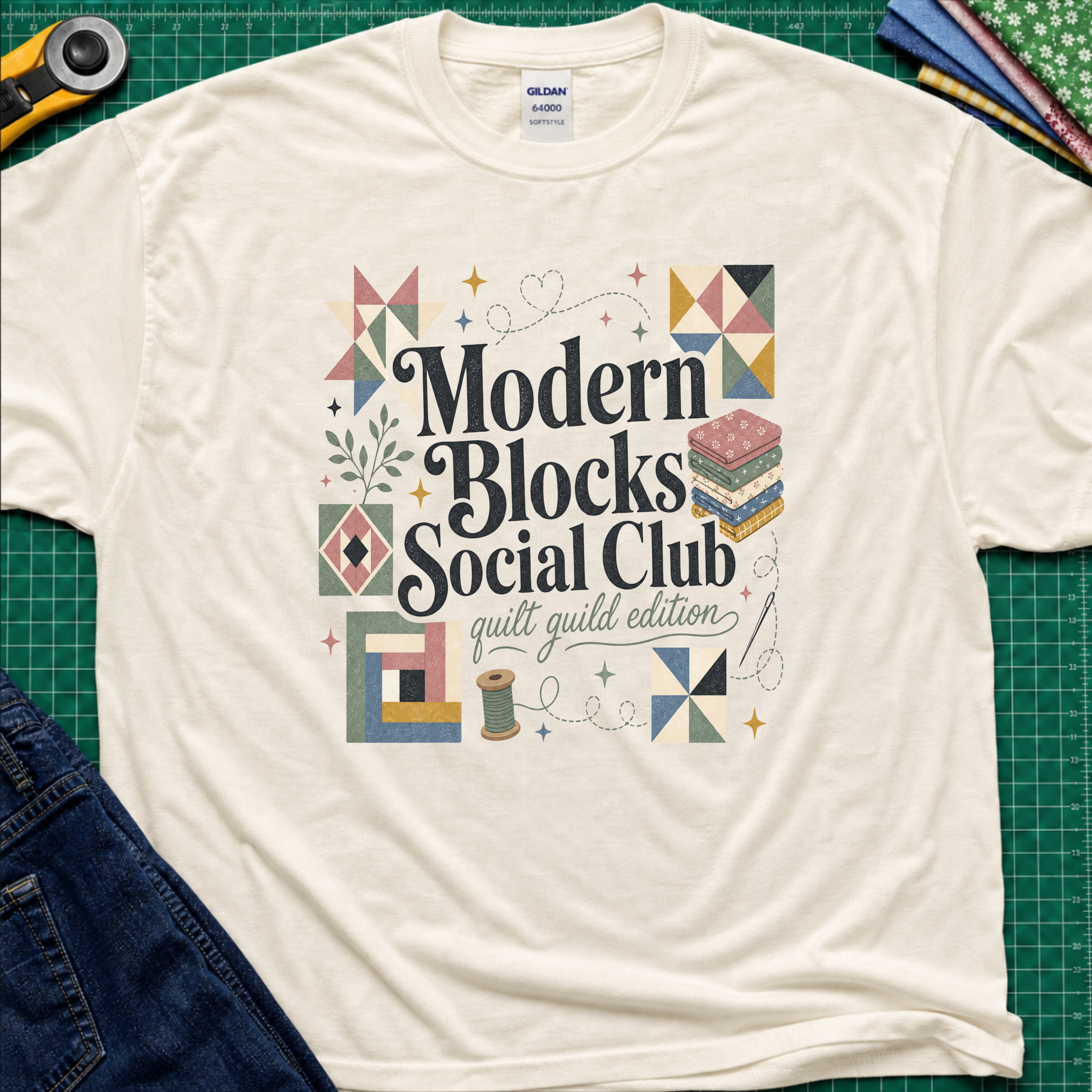 A light cream Gildan t-shirt with a colorful retro-style graphic reading 'Modern Blocks Social Club quilt guild edition' surrounded by quilt block illustrations. The shirt lays flat on a green cutting mat with a rotary cutter, fabric stack and blue jeans partially visible in the corners.
