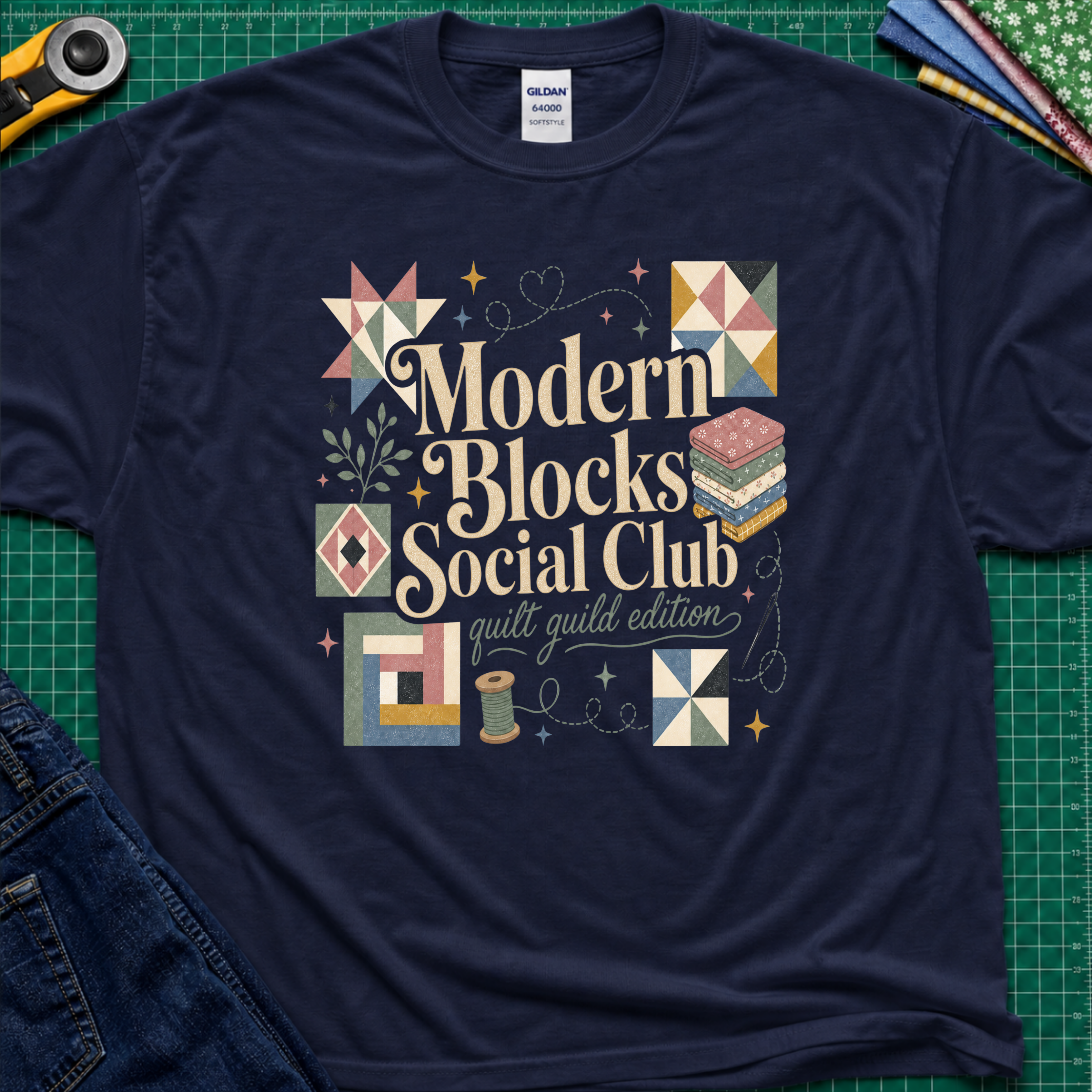 A navy blue Gildan t-shirt with a colorful retro-style graphic reading 'Modern Blocks Social Club quilt guild edition' surrounded by quilt block illustrations. The shirt lays flat on a green cutting mat with a rotary cutter, fabric stack and blue jeans partially visible in the corners.