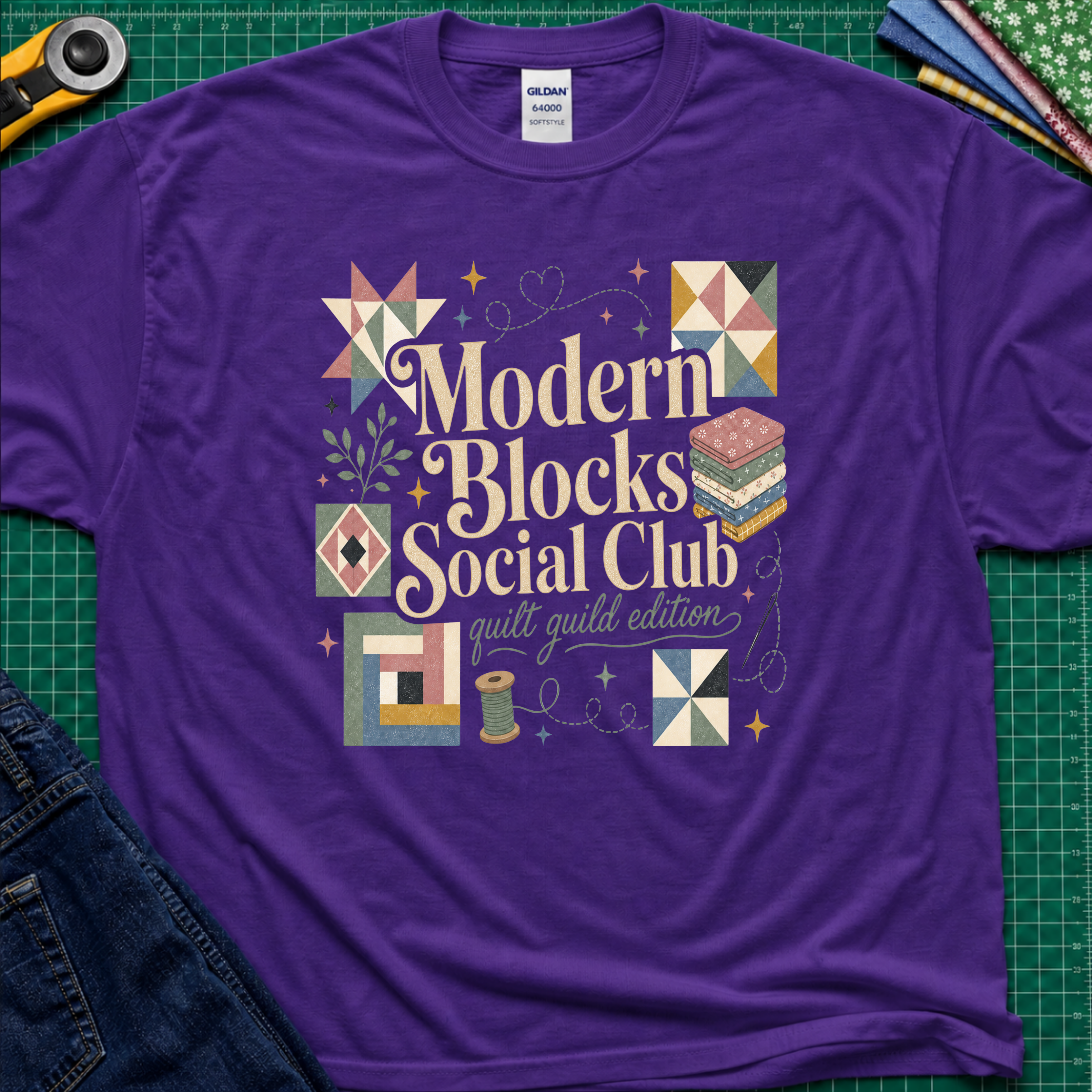 A purple Gildan t-shirt with a colorful retro-style graphic reading 'Modern Blocks Social Club quilt guild edition' surrounded by quilt block illustrations. The shirt lays flat on a green cutting mat with a rotary cutter, fabric stack and blue jeans partially visible in the corners.