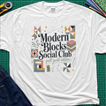 A white Gildan t-shirt with a colorful retro-style graphic reading 'Modern Blocks Social Club quilt guild edition' surrounded by quilt block illustrations. The shirt lays flat on a green cutting mat with a rotary cutter, fabric stack and blue jeans partially visible in the corners.