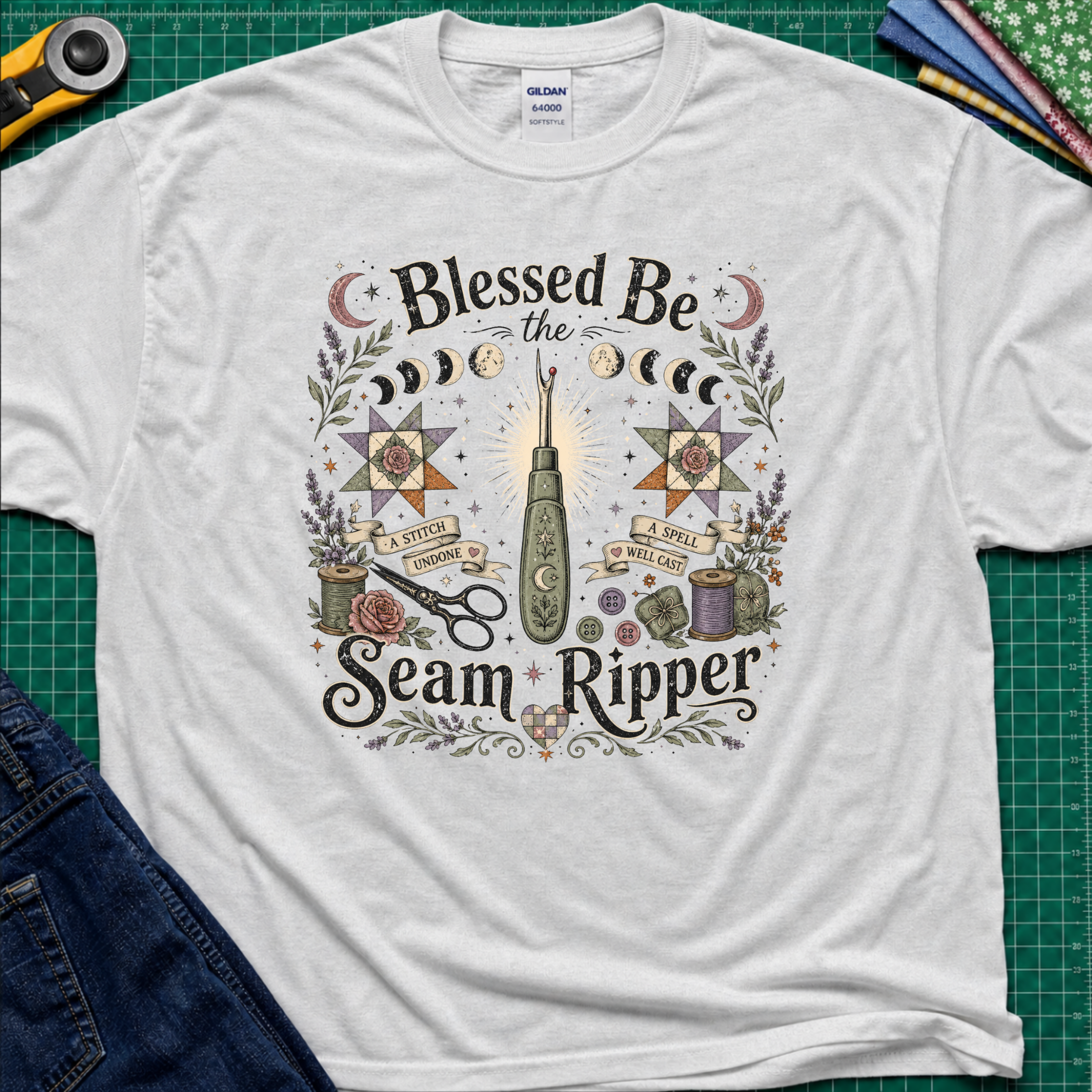 A light grey Gildan t-shirt with a detailed graphic print reading 'Blessed Be the Seam Ripper' surrounded by moon phases, quilt stars, thread spools with a back lit seam ripper in the middle. The t-shirt lays flat on a green cutting mat with a rotary cutter, folded fabric stack and blue jeans partially visible in the corners.