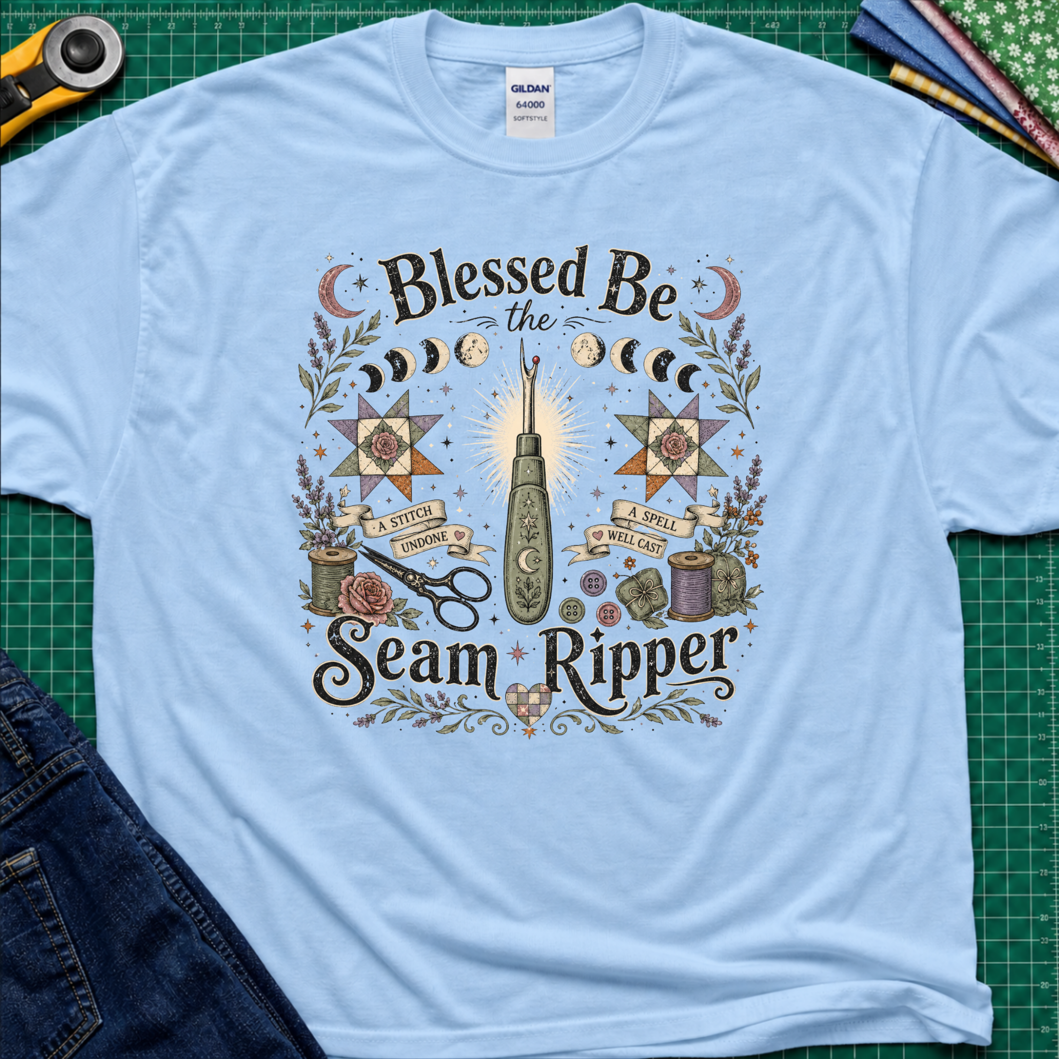 A light blue Gildan t-shirt with a detailed graphic print reading 'Blessed Be the Seam Ripper' surrounded by moon phases, quilt stars, thread spools with a back lit seam ripper in the middle. The t-shirt lays flat on a green cutting mat with a rotary cutter, folded fabric stack and blue jeans partially visible in the corners.