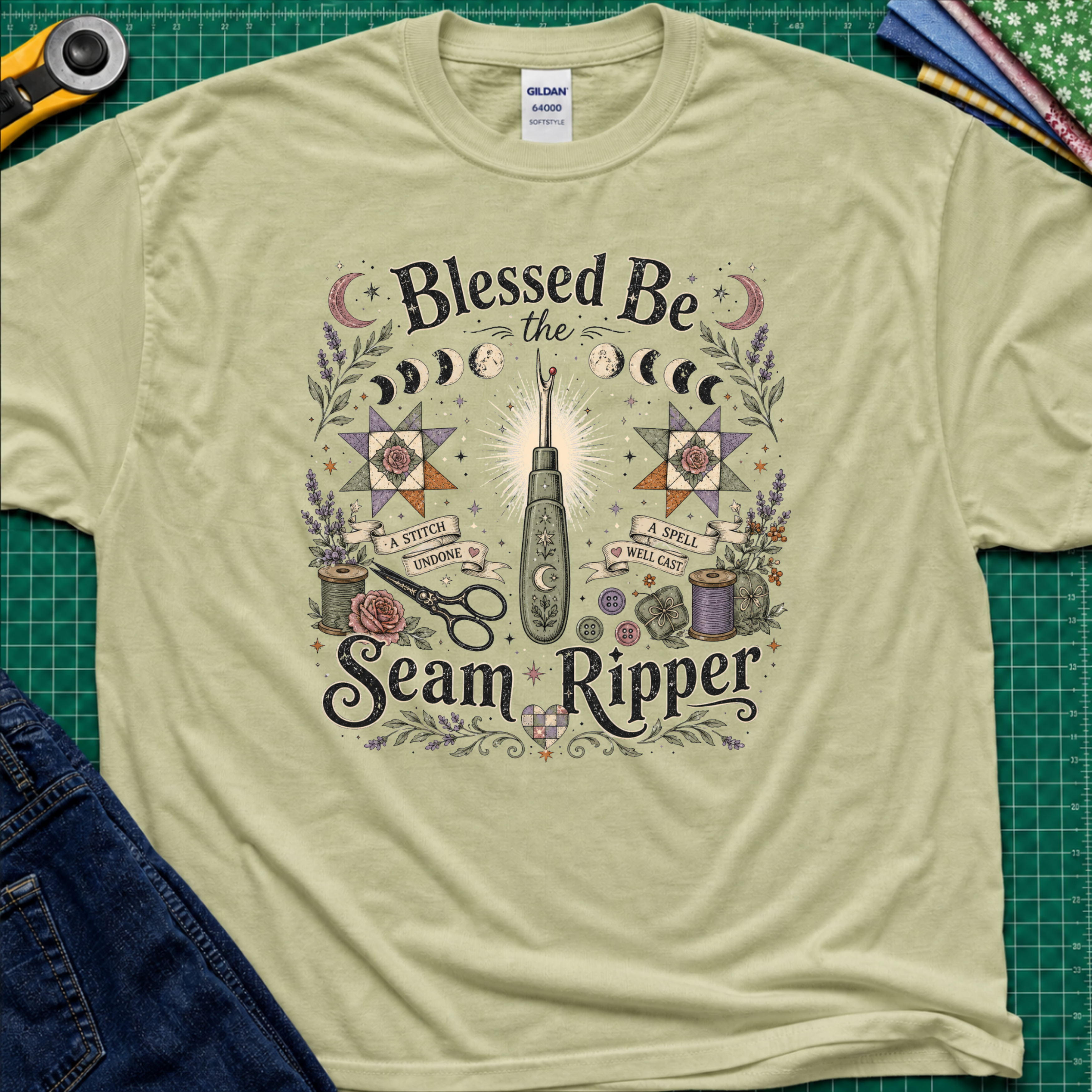 A light pistachio green Gildan t-shirt with a detailed graphic print reading 'Blessed Be the Seam Ripper' surrounded by moon phases, quilt stars, thread spools with a back lit seam ripper in the middle. The t-shirt lays flat on a green cutting mat with a rotary cutter, folded fabric stack and blue jeans partially visible in the corners.