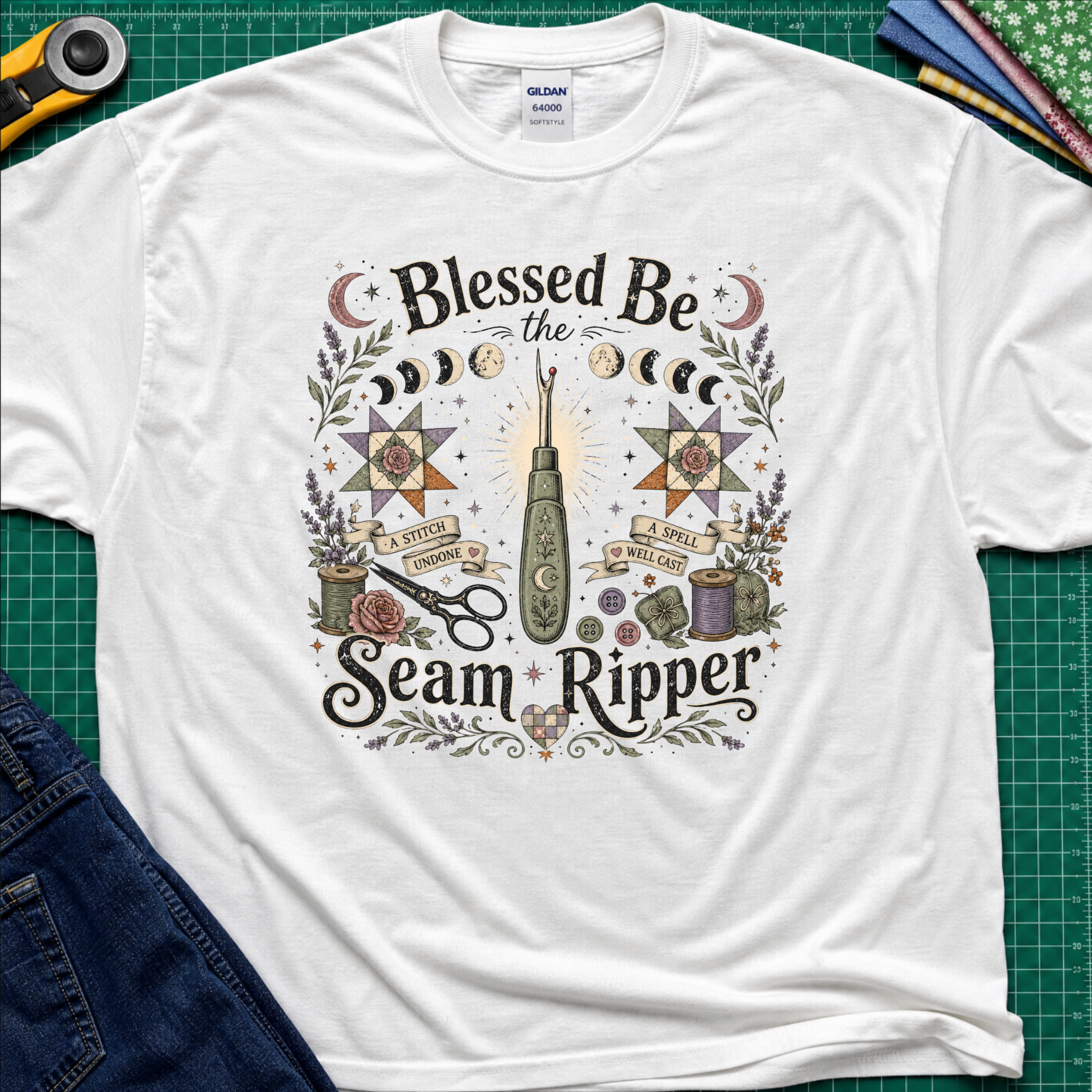 A white Gildan t-shirt with a detailed graphic print reading 'Blessed Be the Seam Ripper' surrounded by moon phases, quilt stars, thread spools with a back lit seam ripper in the middle. The t-shirt lays flat on a green cutting mat with a rotary cutter, folded fabric stack and blue jeans partially visible in the corners.