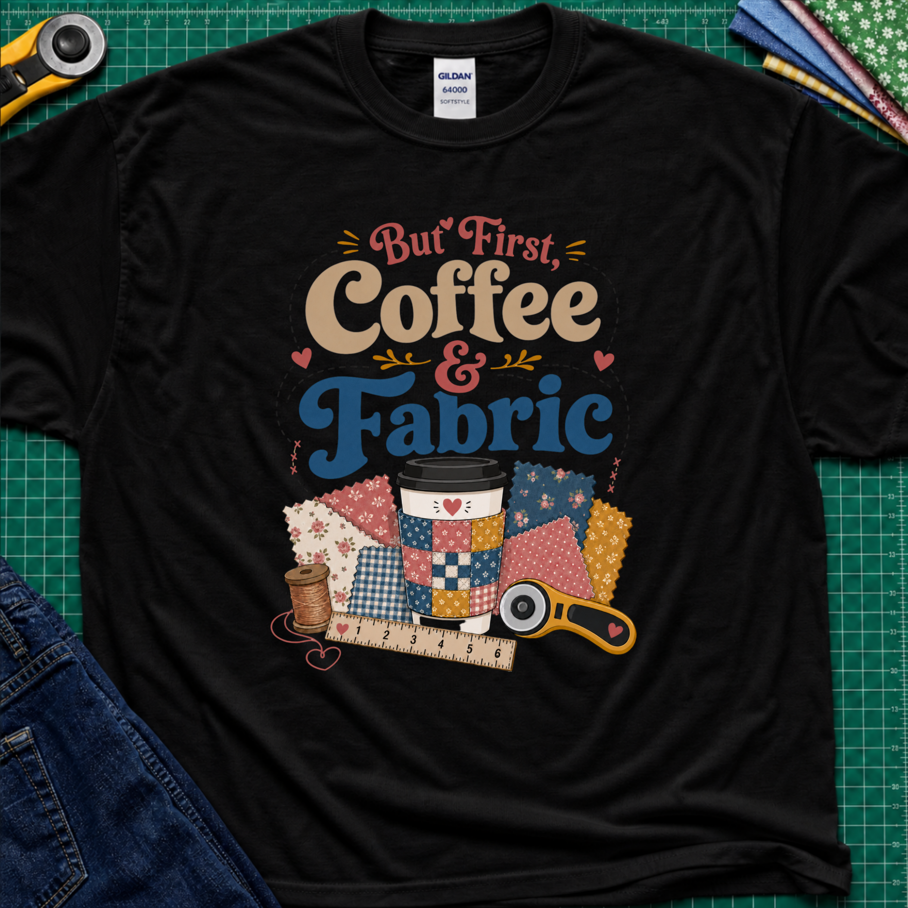 A black Gildan t-shirt with a colorful graphic that reads 'But First, Coffee & Fabric' above a quilted coffee cup, fabric swatches, thread spool, ruler, and rotary cutter. The shirt is laid flat on a green cutting mat with sewing supplies and blue jeans partially visible around the edges.