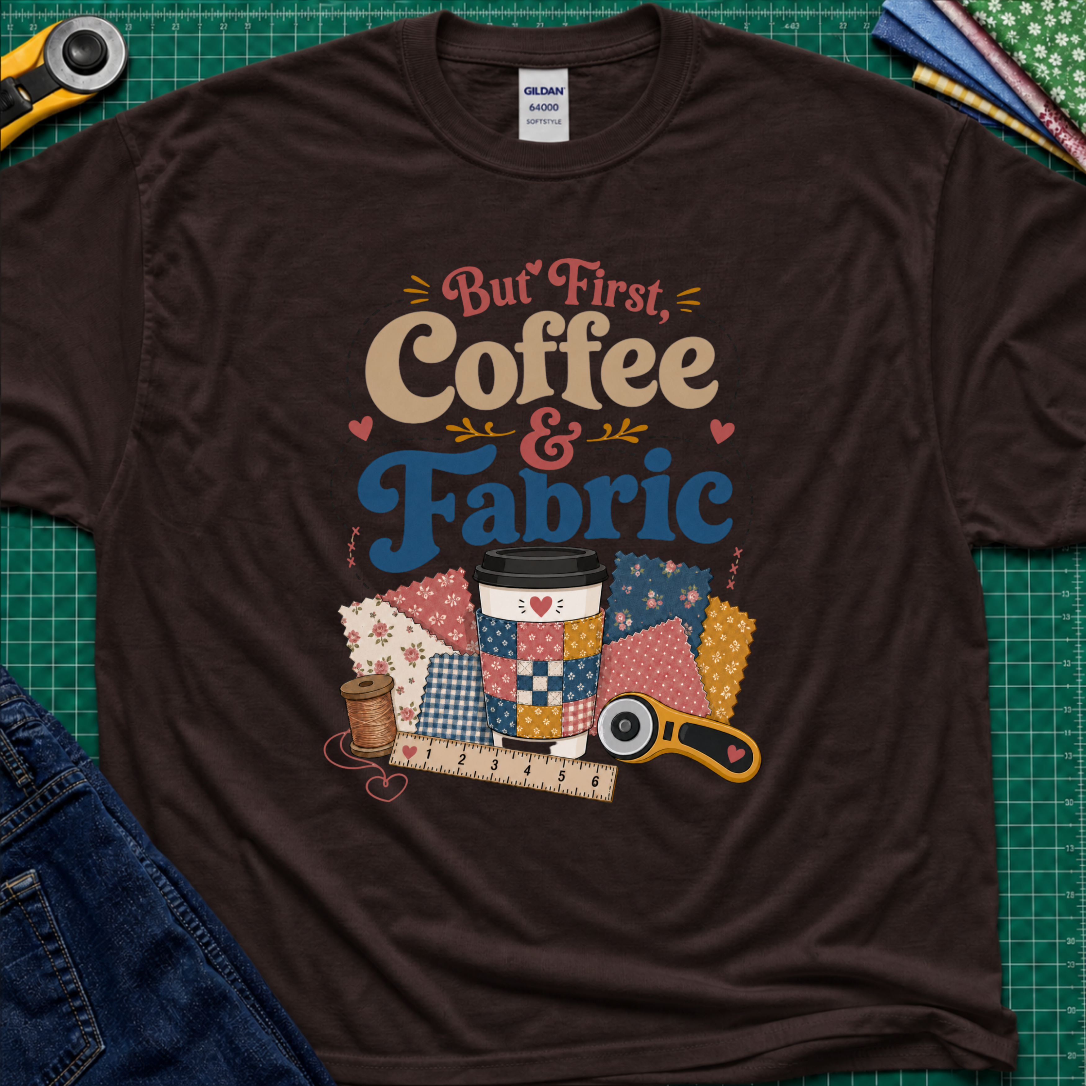 A chocolate brown Gildan t-shirt with a colorful graphic that reads 'But First, Coffee & Fabric' above a quilted coffee cup, fabric swatches, thread spool, ruler, and rotary cutter. The shirt is laid flat on a green cutting mat with sewing supplies and blue jeans partially visible around the edges.