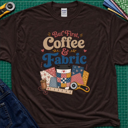 A chocolate brown Gildan t-shirt with a colorful graphic that reads 'But First, Coffee & Fabric' above a quilted coffee cup, fabric swatches, thread spool, ruler, and rotary cutter. The shirt is laid flat on a green cutting mat with sewing supplies and blue jeans partially visible around the edges.