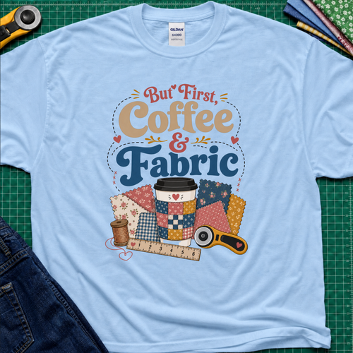 A light blue Gildan t-shirt with a colorful graphic that reads 'But First, Coffee & Fabric' above a quilted coffee cup, fabric swatches, thread spool, ruler, and rotary cutter. The shirt is laid flat on a green cutting mat with sewing supplies and blue jeans partially visible around the edges.