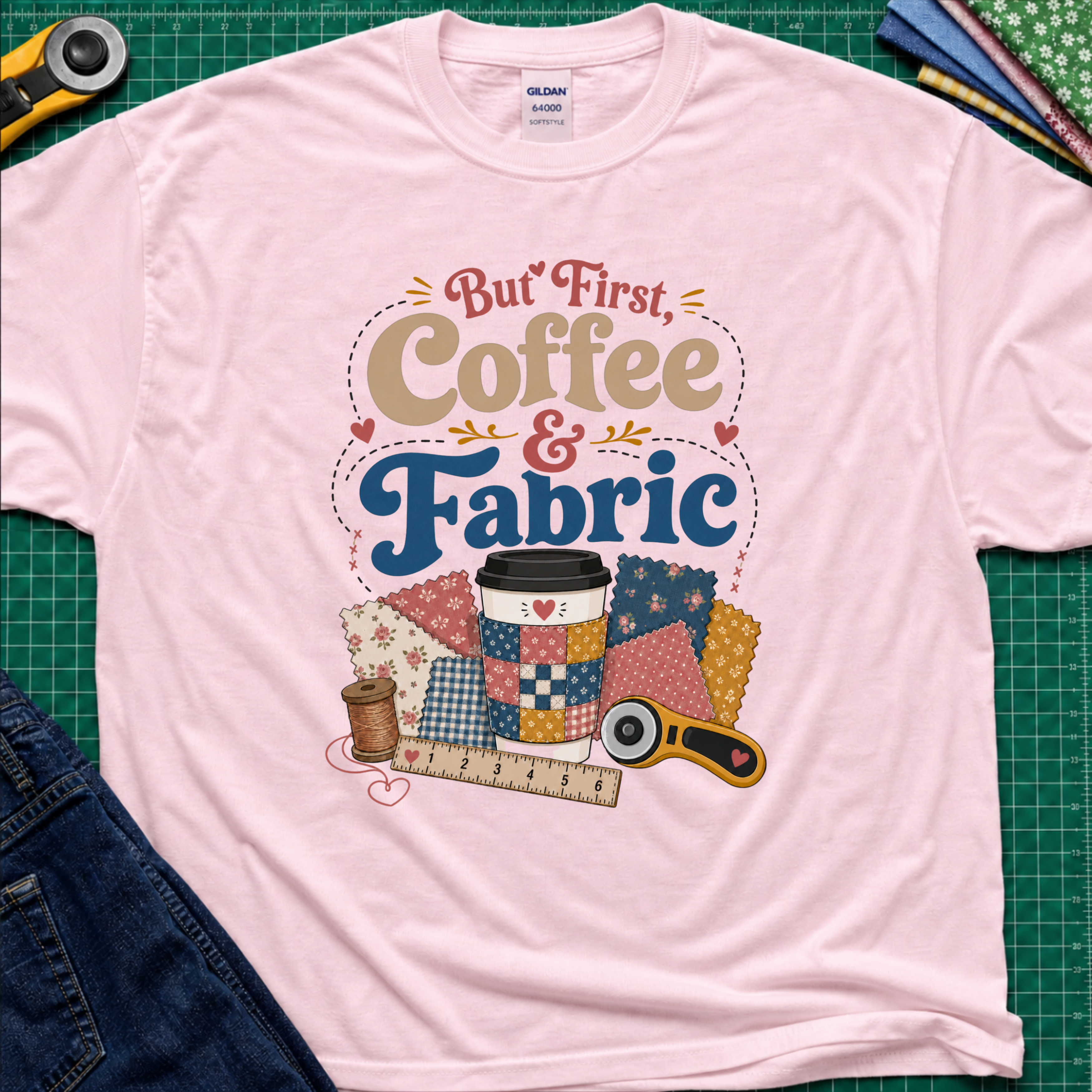 A light pink Gildan t-shirt with a colorful graphic that reads 'But First, Coffee & Fabric' above a quilted coffee cup, fabric swatches, thread spool, ruler, and rotary cutter. The shirt is laid flat on a green cutting mat with sewing supplies and blue jeans partially visible around the edges.