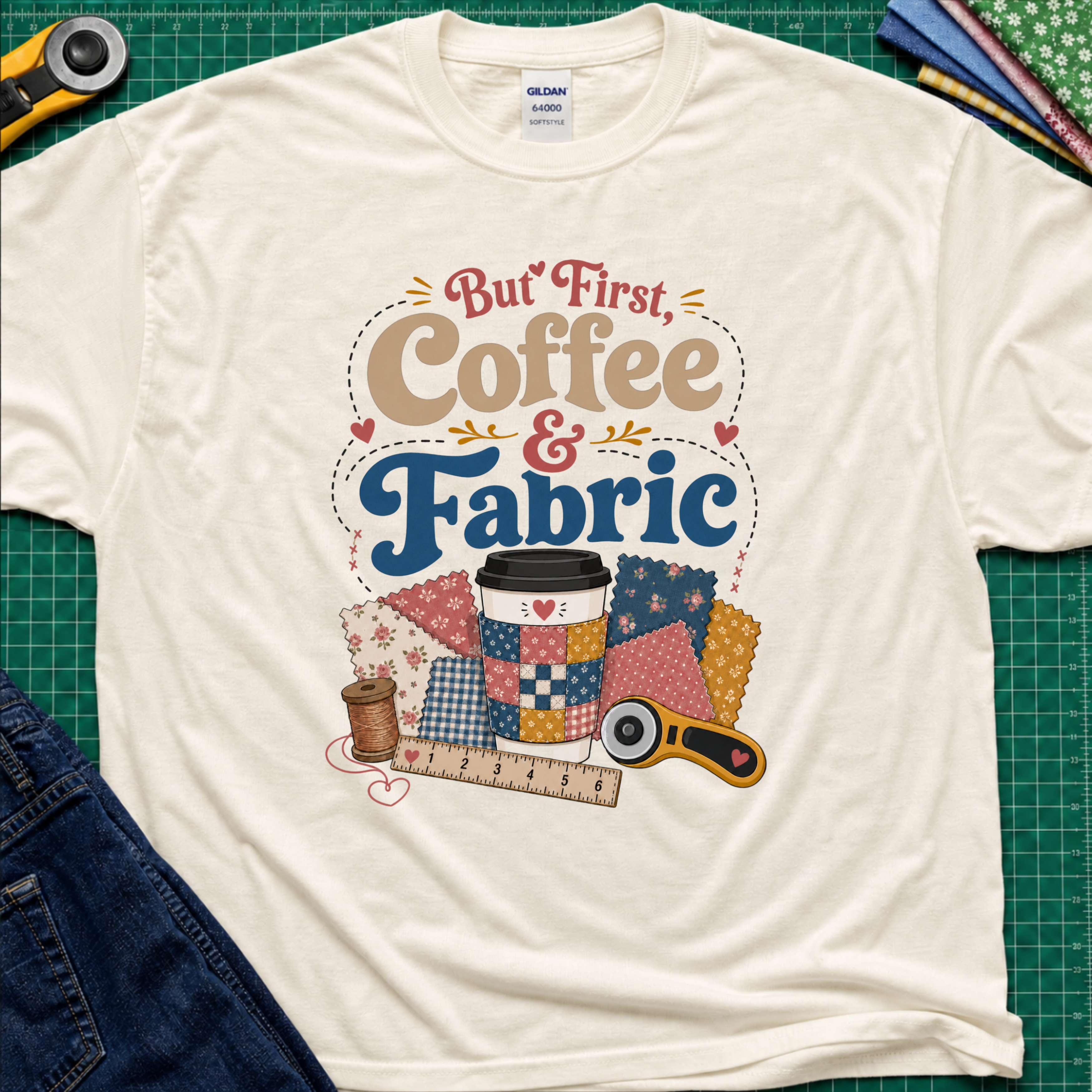 A light cream Gildan t-shirt with a colorful graphic that reads 'But First, Coffee & Fabric' above a quilted coffee cup, fabric swatches, thread spool, ruler, and rotary cutter. The shirt is laid flat on a green cutting mat with sewing supplies and blue jeans partially visible around the edges.