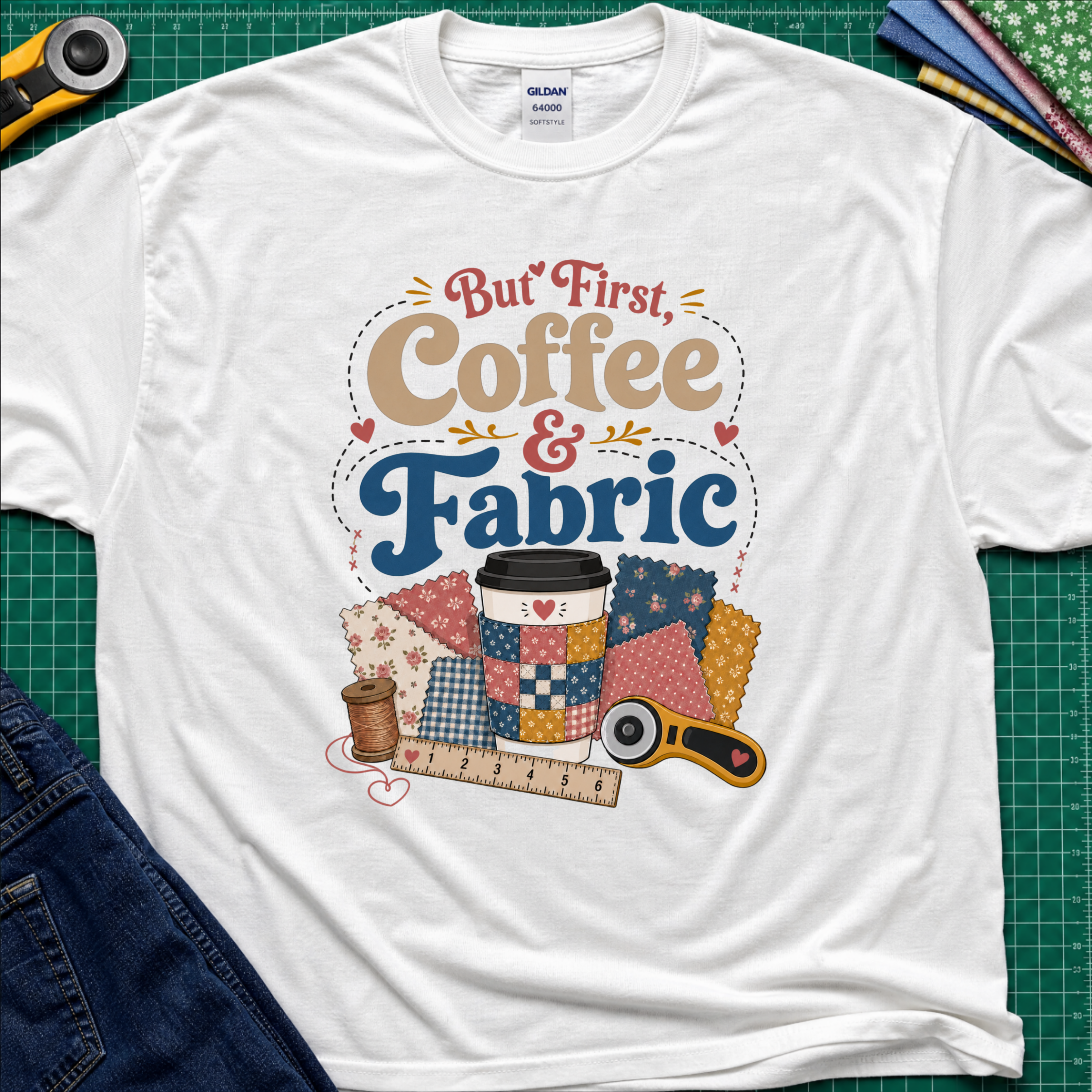 A white Gildan t-shirt with a colorful graphic that reads 'But First, Coffee & Fabric' above a quilted coffee cup, fabric swatches, thread spool, ruler, and rotary cutter. The shirt is laid flat on a green cutting mat with sewing supplies and blue jeans partially visible around the edges.
