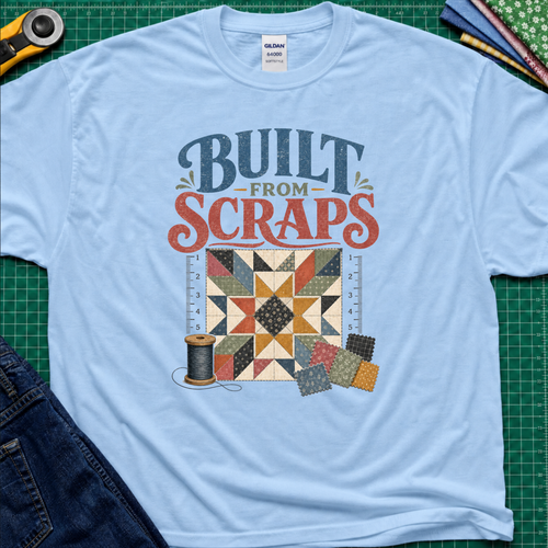 A light blue Gildan t-shirt with a retro-style quilt graphic and the text 'BUILT FROM SCRAPS' in blue and coral. The shirt is styled in a flat-lay on a green cutting mat with sewing tools and fabric.