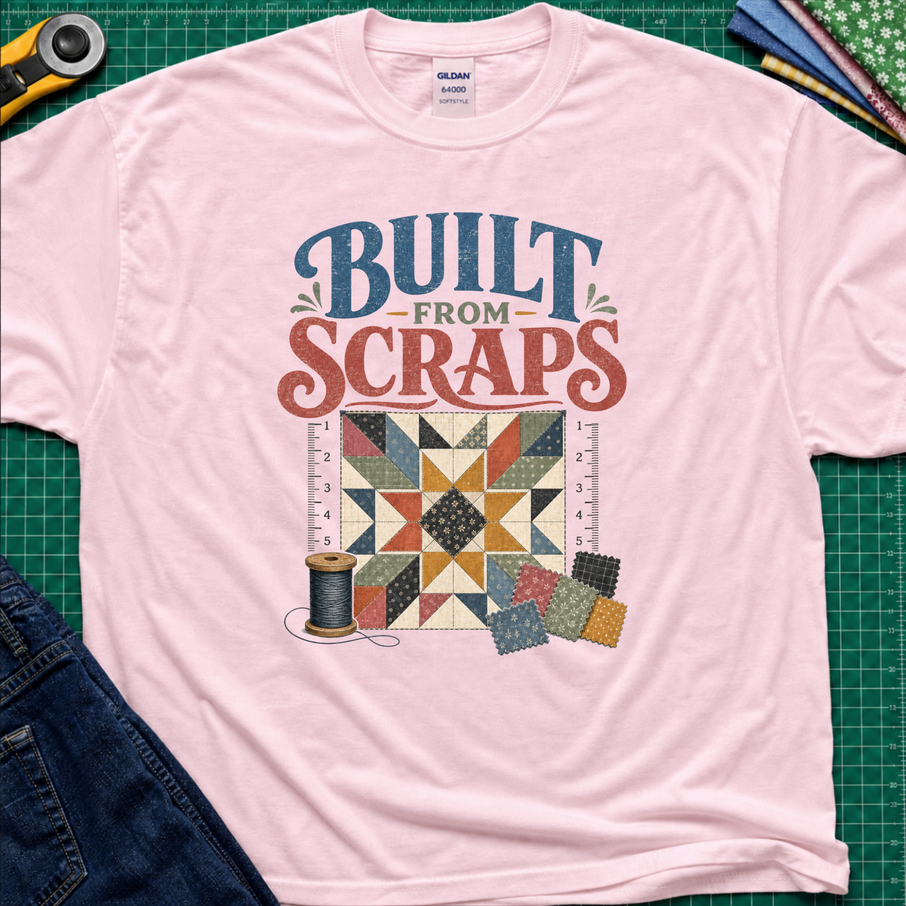 A light pink Gildan t-shirt with a retro-style quilt graphic and the text 'BUILT FROM SCRAPS' in blue and coral. The shirt is styled in a flat-lay on a green cutting mat with sewing tools and fabric.