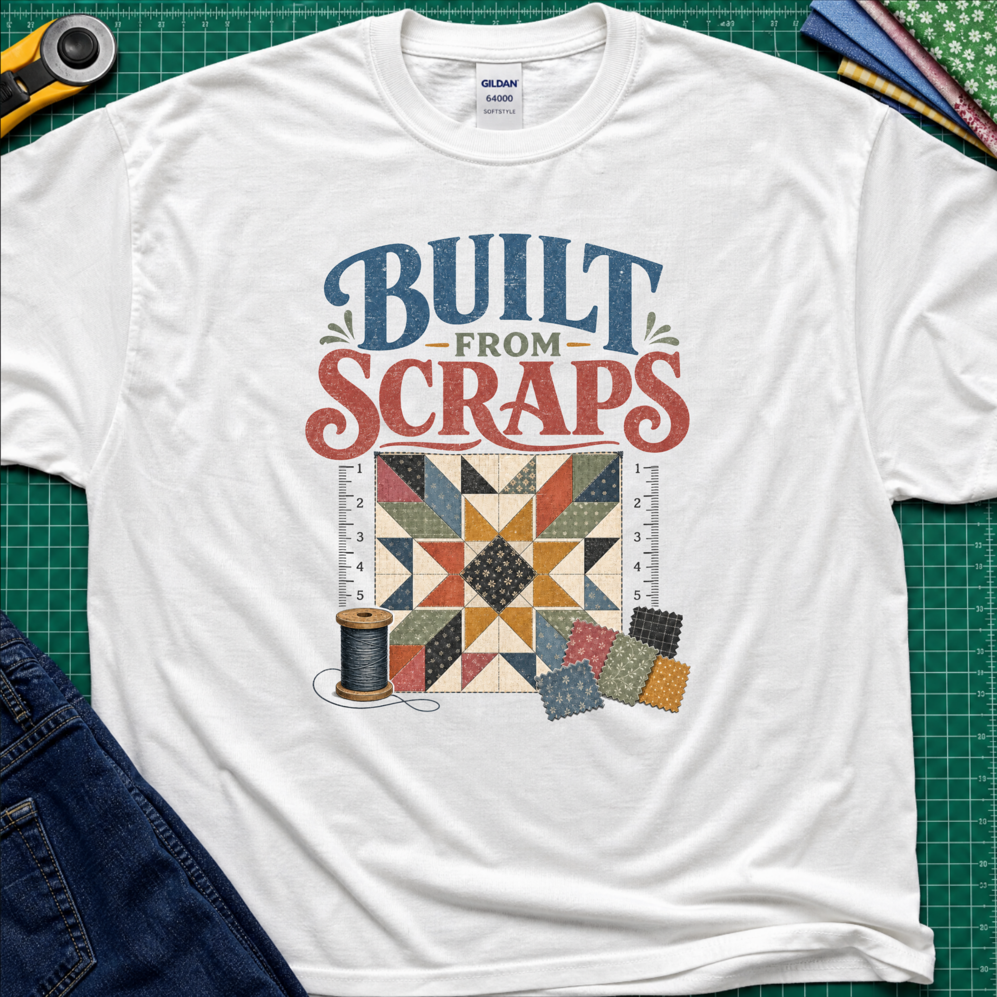 A white Gildan t-shirt with a retro-style quilt graphic and the text 'BUILT FROM SCRAPS' in blue and coral. The shirt is styled in a flat-lay on a green cutting mat with sewing tools and fabric.