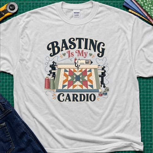 A light grey Gildan t-shirt with a graphic print that reads 'BASTING IS MY CARDIO' around an illustration of a longarm machine and quilt. The shirt is laid flat on a green cutting mat with craft tools and jeans partially visible around it.