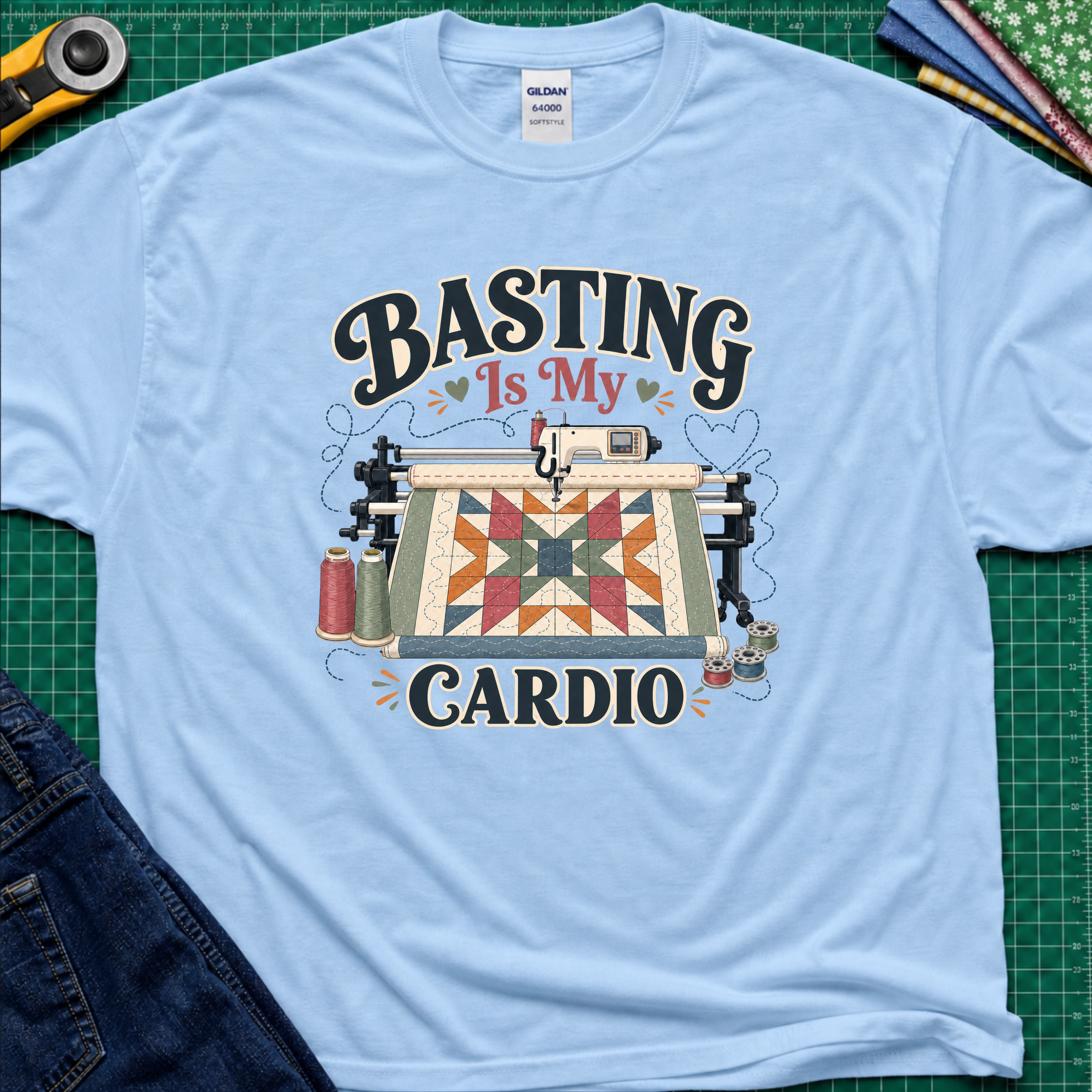 A light blue Gildan t-shirt with a graphic print that reads 'BASTING IS MY CARDIO' around an illustration of a longarm machine and quilt. The shirt is laid flat on a green cutting mat with craft tools and jeans partially visible around it.