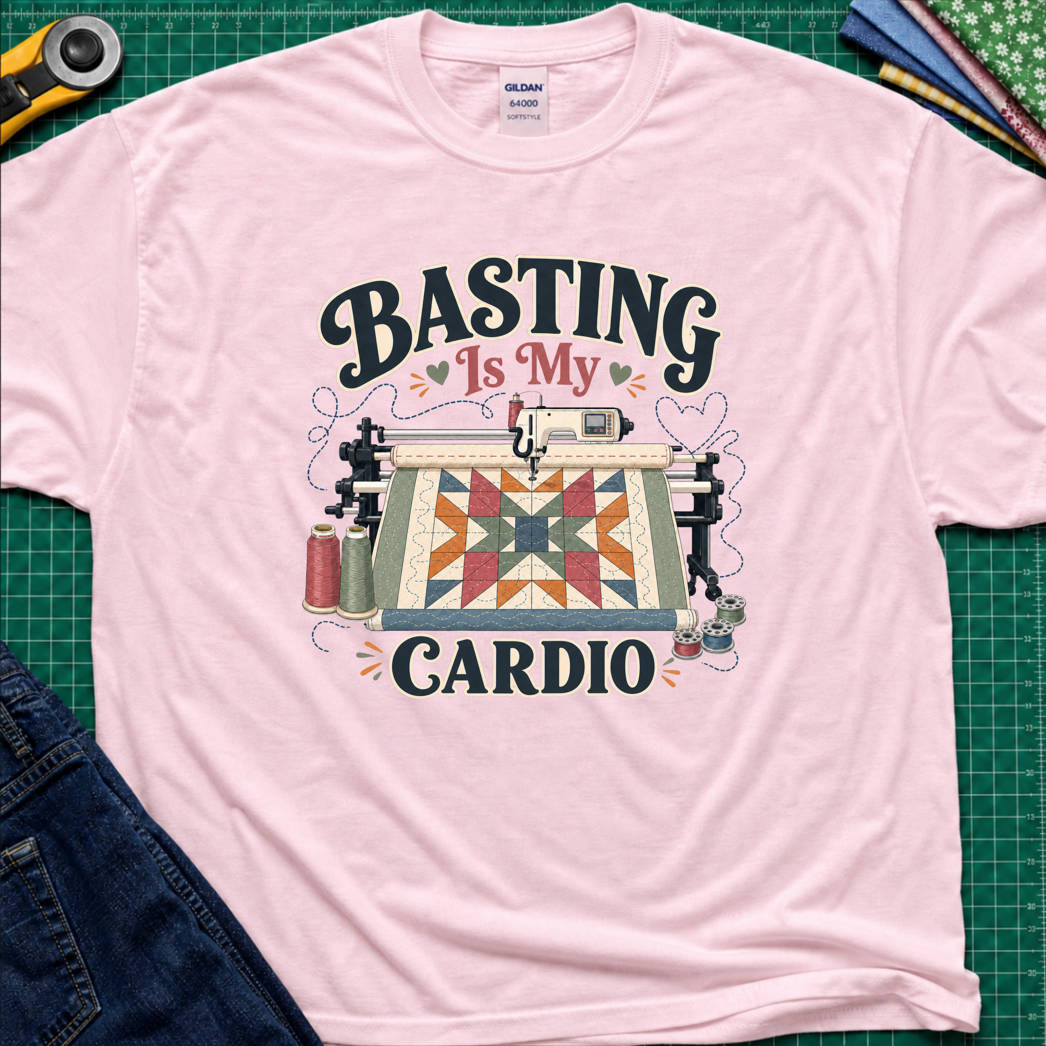 A light pink Gildan t-shirt with a graphic print that reads 'BASTING IS MY CARDIO' around an illustration of a longarm machine and quilt. The shirt is laid flat on a green cutting mat with craft tools and jeans partially visible around it.