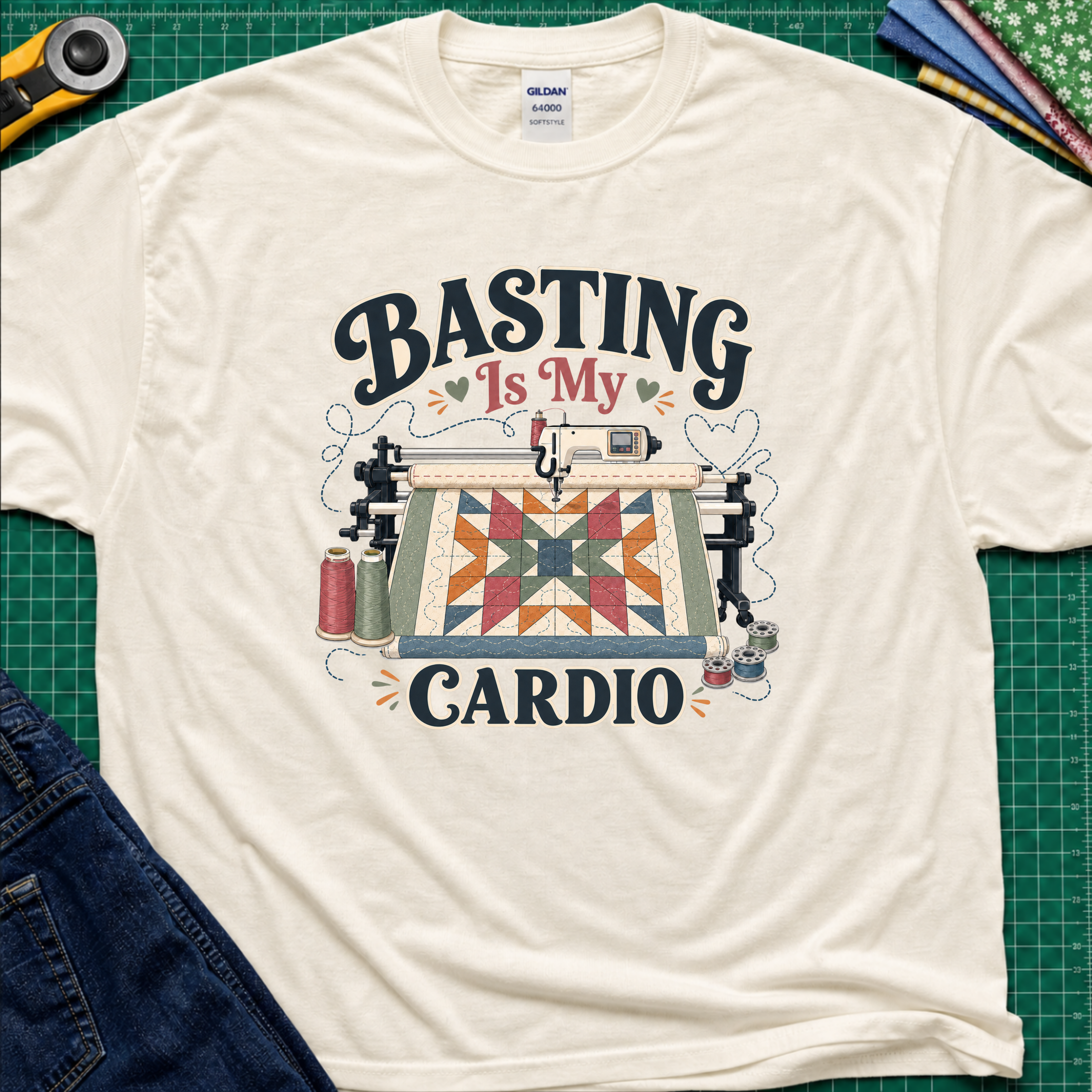 A light cream Gildan t-shirt with a graphic print that reads 'BASTING IS MY CARDIO' around an illustration of a longarm machine and quilt. The shirt is laid flat on a green cutting mat with craft tools and jeans partially visible around it.