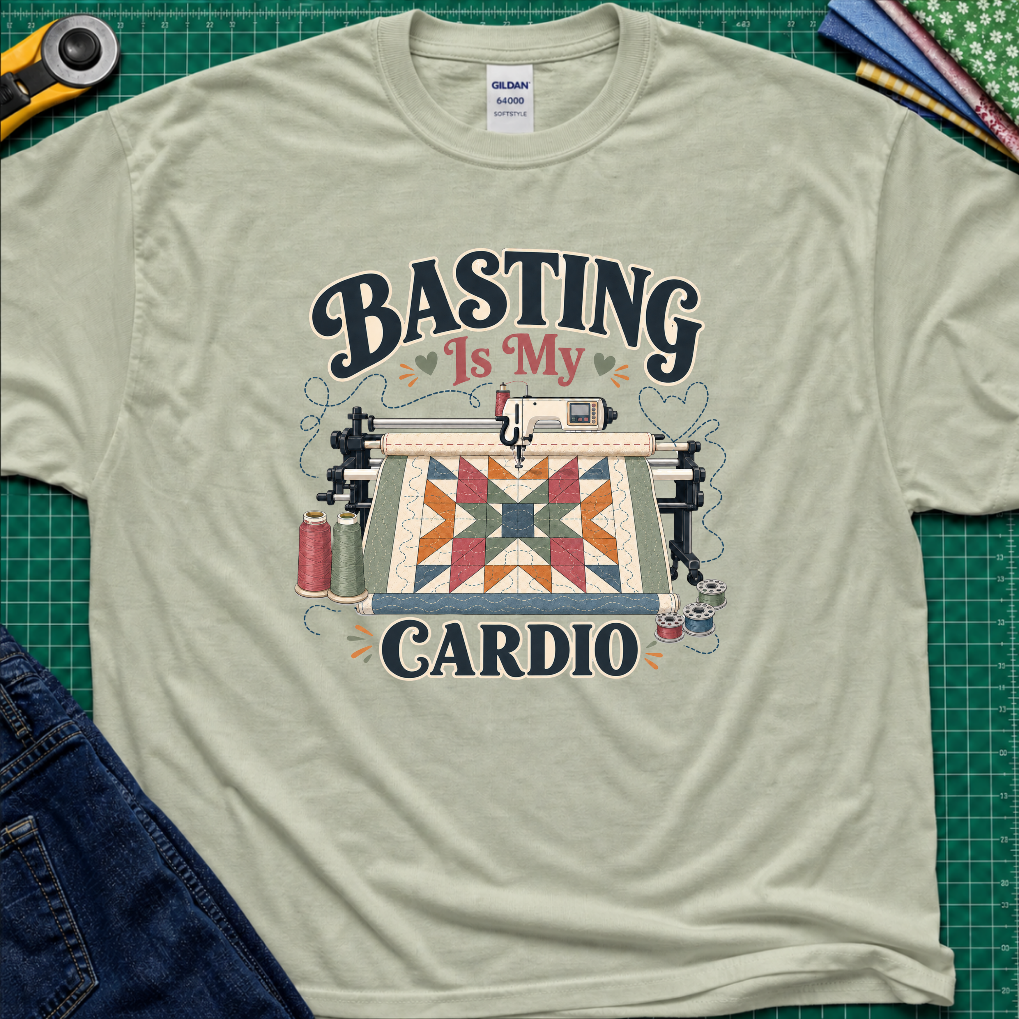 A light sage green Gildan t-shirt with a graphic print that reads 'BASTING IS MY CARDIO' around an illustration of a longarm machine and quilt. The shirt is laid flat on a green cutting mat with craft tools and jeans partially visible around it.