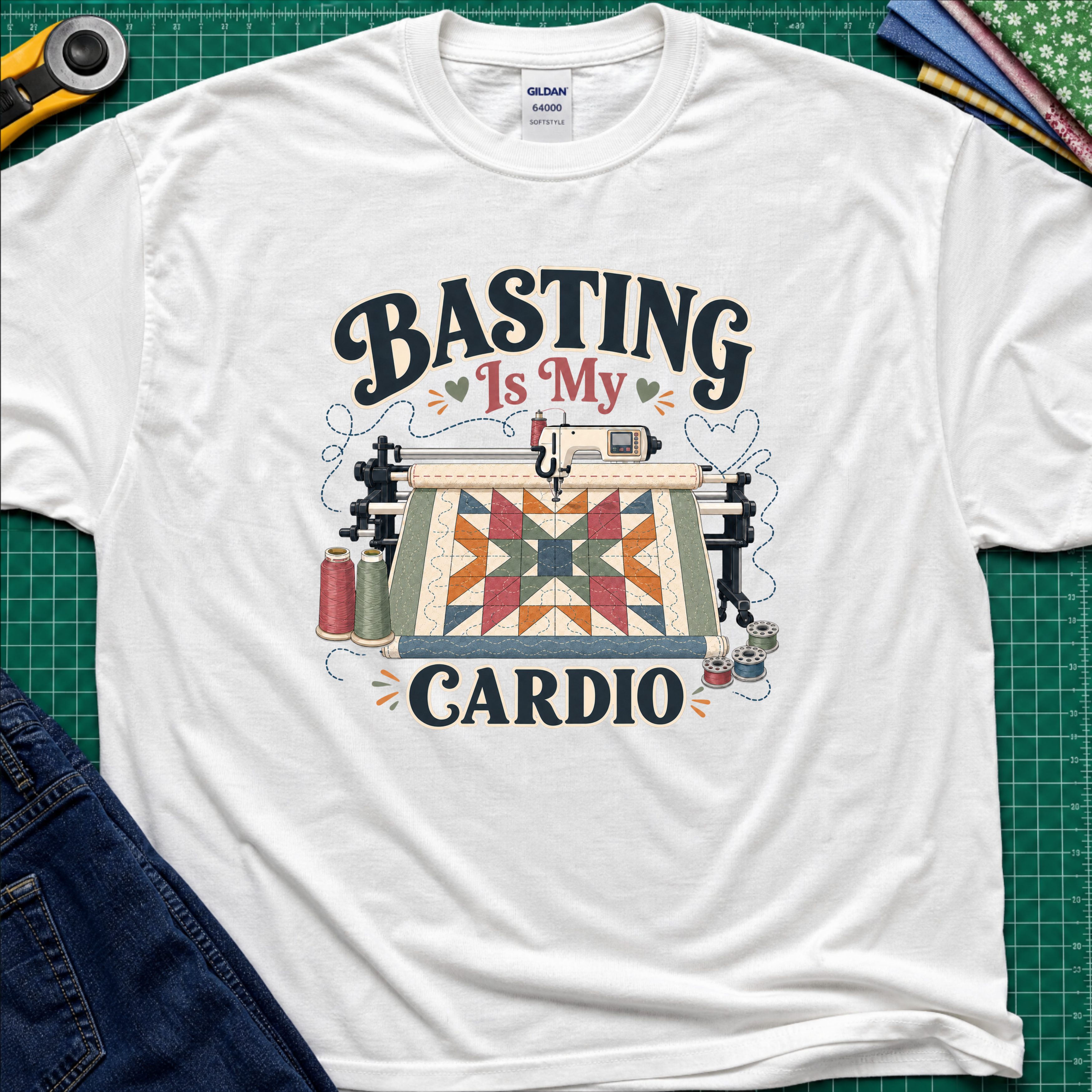 A white Gildan t-shirt with a graphic print that reads 'BASTING IS MY CARDIO' around an illustration of a longarm machine and quilt. The shirt is laid flat on a green cutting mat with craft tools and jeans partially visible around it.