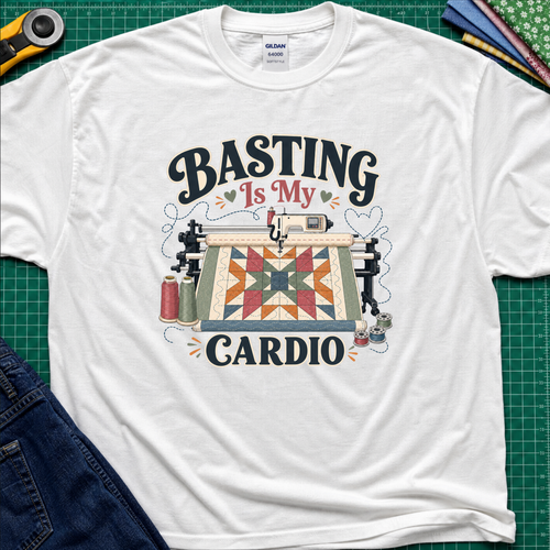 A white Gildan t-shirt with a graphic print that reads 'BASTING IS MY CARDIO' around an illustration of a longarm machine and quilt. The shirt is laid flat on a green cutting mat with craft tools and jeans partially visible around it.