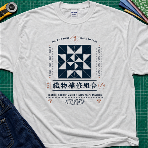A light grey Gildan t-shirt with a central graphic of a quilt block and Japanese text reading 'Textile Repair Guild / Slow Work Division'. The shirt is laid flat on a green cutting mat with a rotary cutter, fabric stack and blue jeans partially visible in the corners.