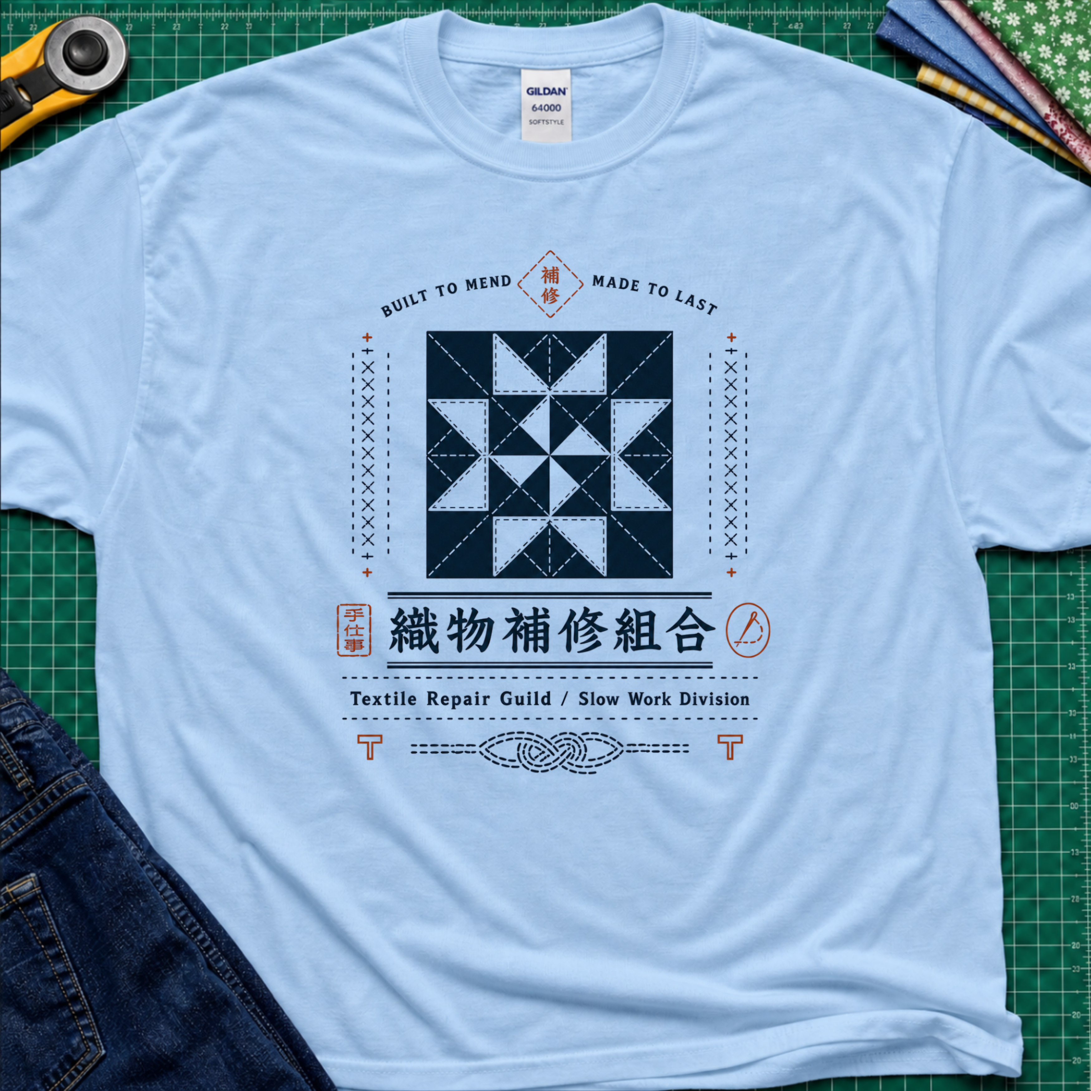 A light blue Gildan t-shirt with a central graphic of a quilt block and Japanese text reading 'Textile Repair Guild / Slow Work Division'. The shirt is laid flat on a green cutting mat with a rotary cutter, fabric stack and blue jeans partially visible in the corners.