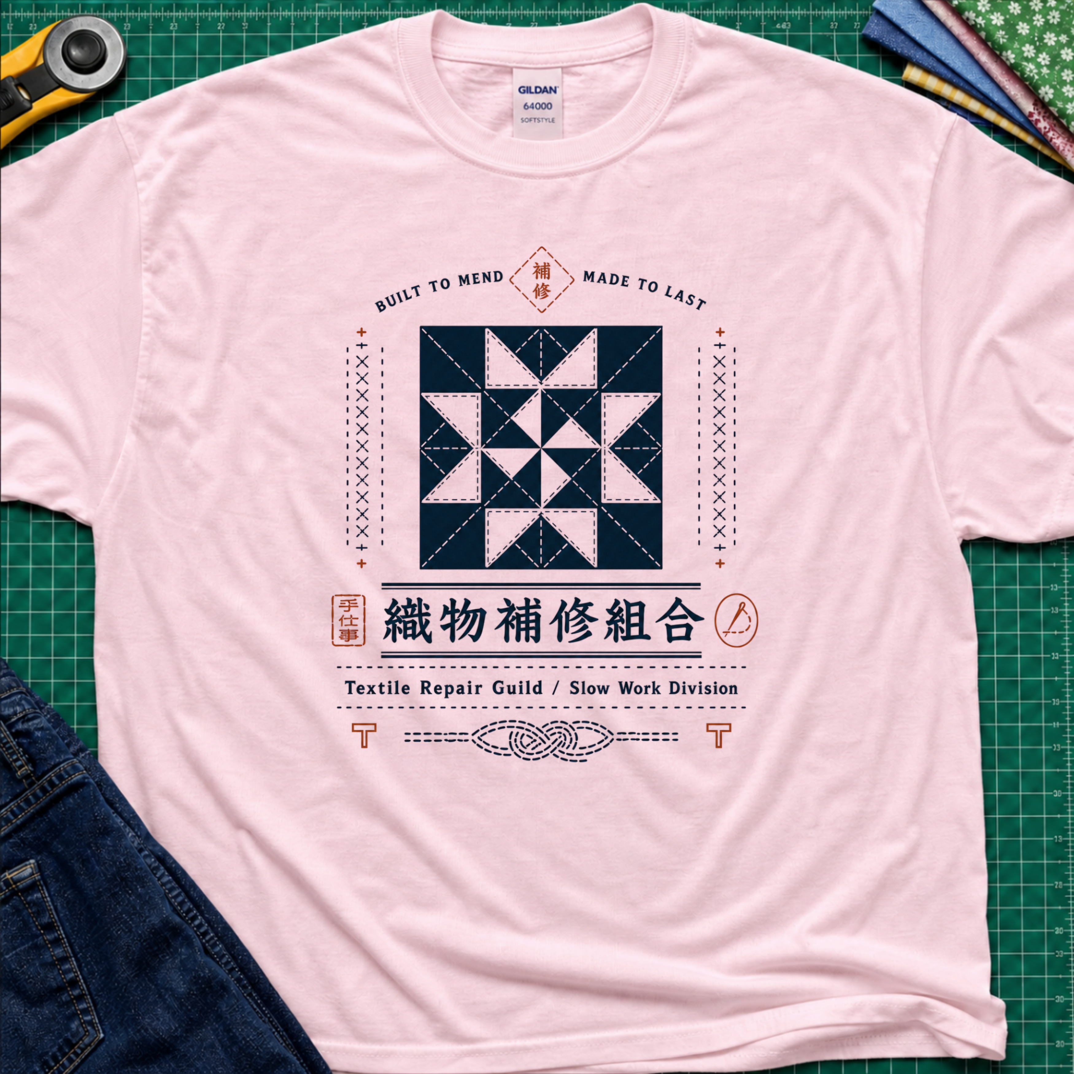 A light pink Gildan t-shirt with a central graphic of a quilt block and Japanese text reading 'Textile Repair Guild / Slow Work Division'. The shirt is laid flat on a green cutting mat with a rotary cutter, fabric stack and blue jeans partially visible in the corners.